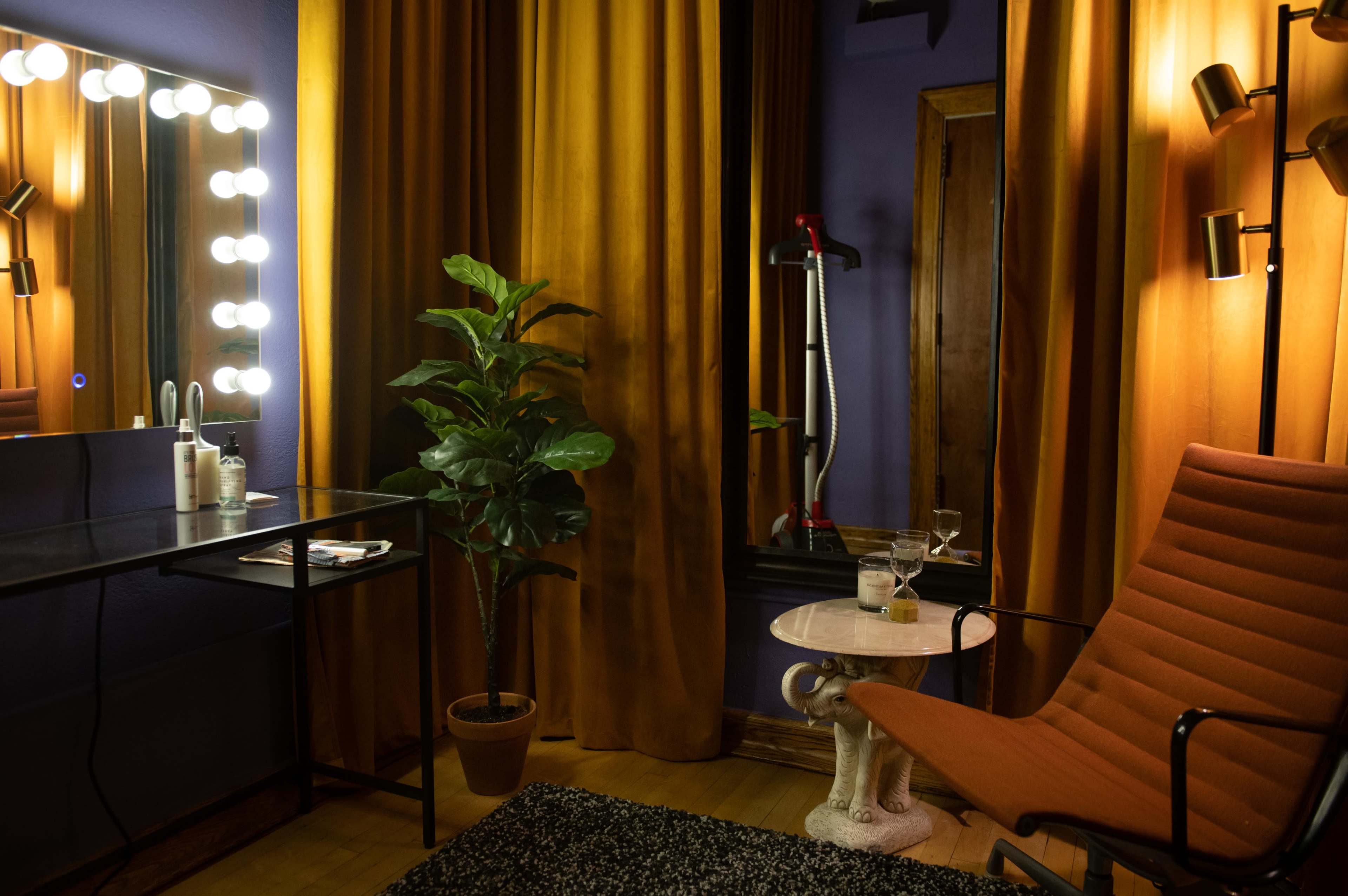 The scene features a cozy room with a vanity mirror, a potted plant, a small side table with a drink, an orange armchair, and curtains in warm tones.