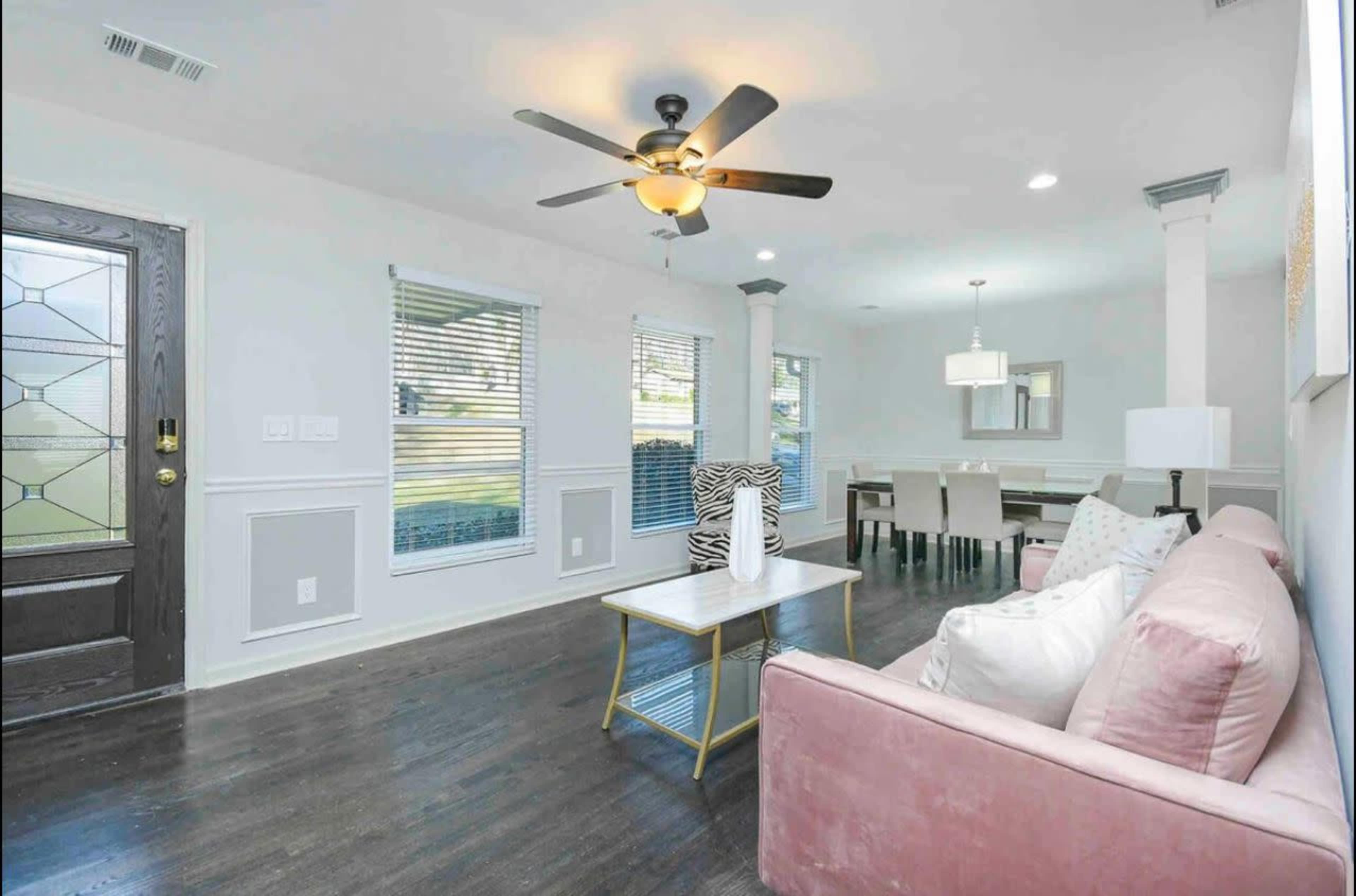 The image shows a modern living room with a pink sofa, a coffee table, a ceiling fan, and a dining area visible in the background.