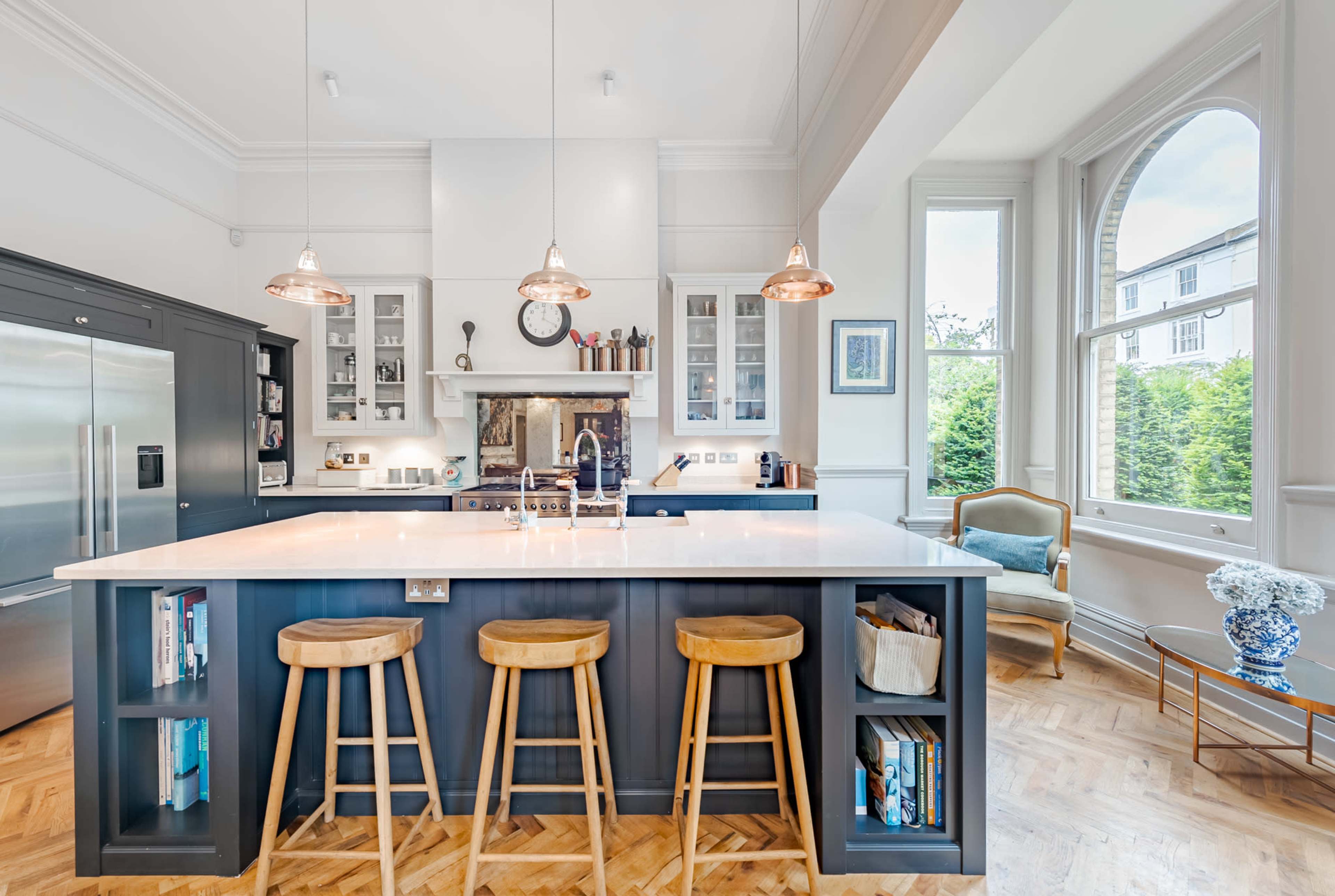 The image shows a modern kitchen with a large island, three wooden barstools, stainless steel appliances, and large windows allowing natural light to flow in.