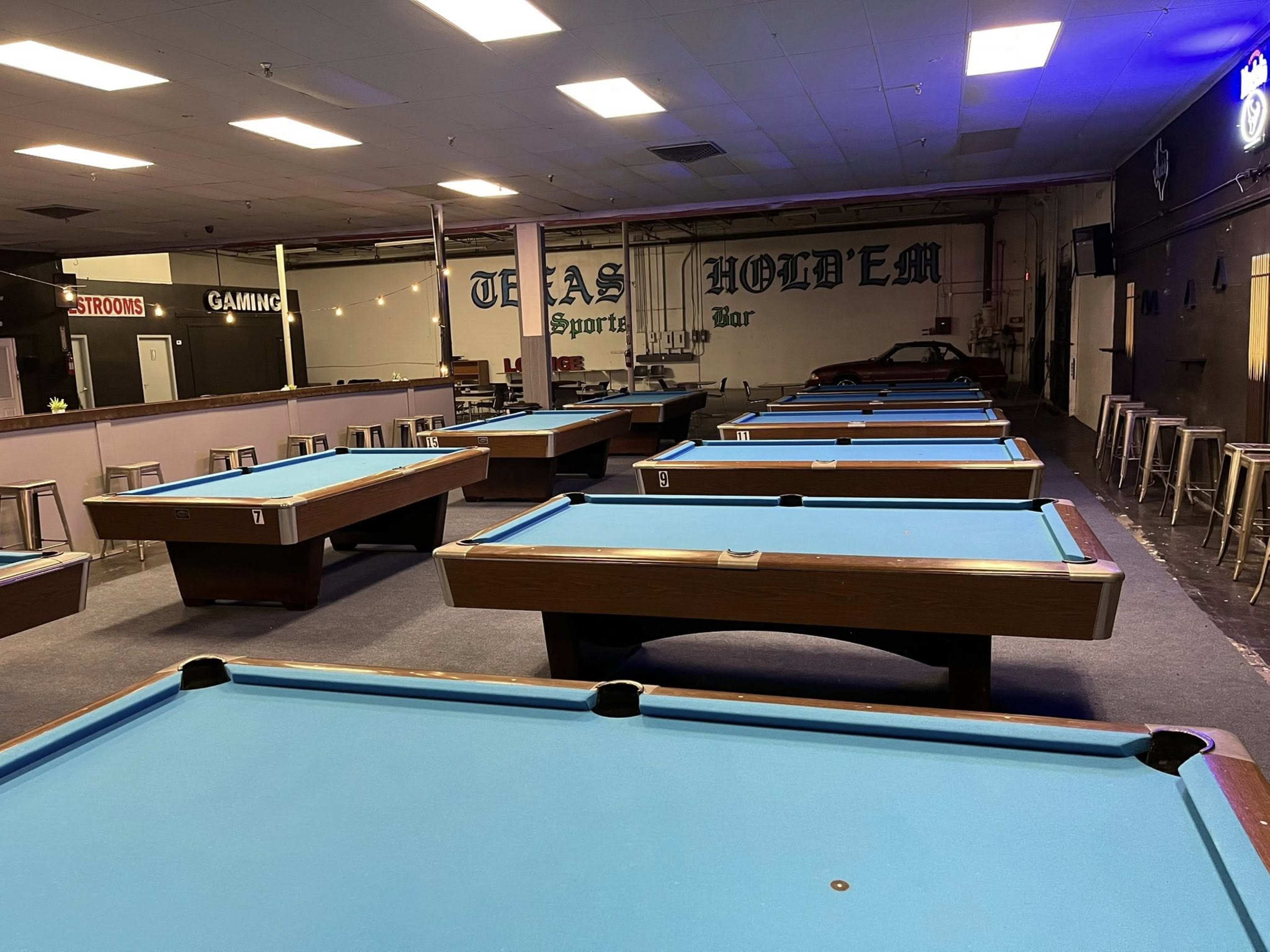 The image shows a spacious billiards hall with multiple pool tables arranged in rows and a bar area in the background.