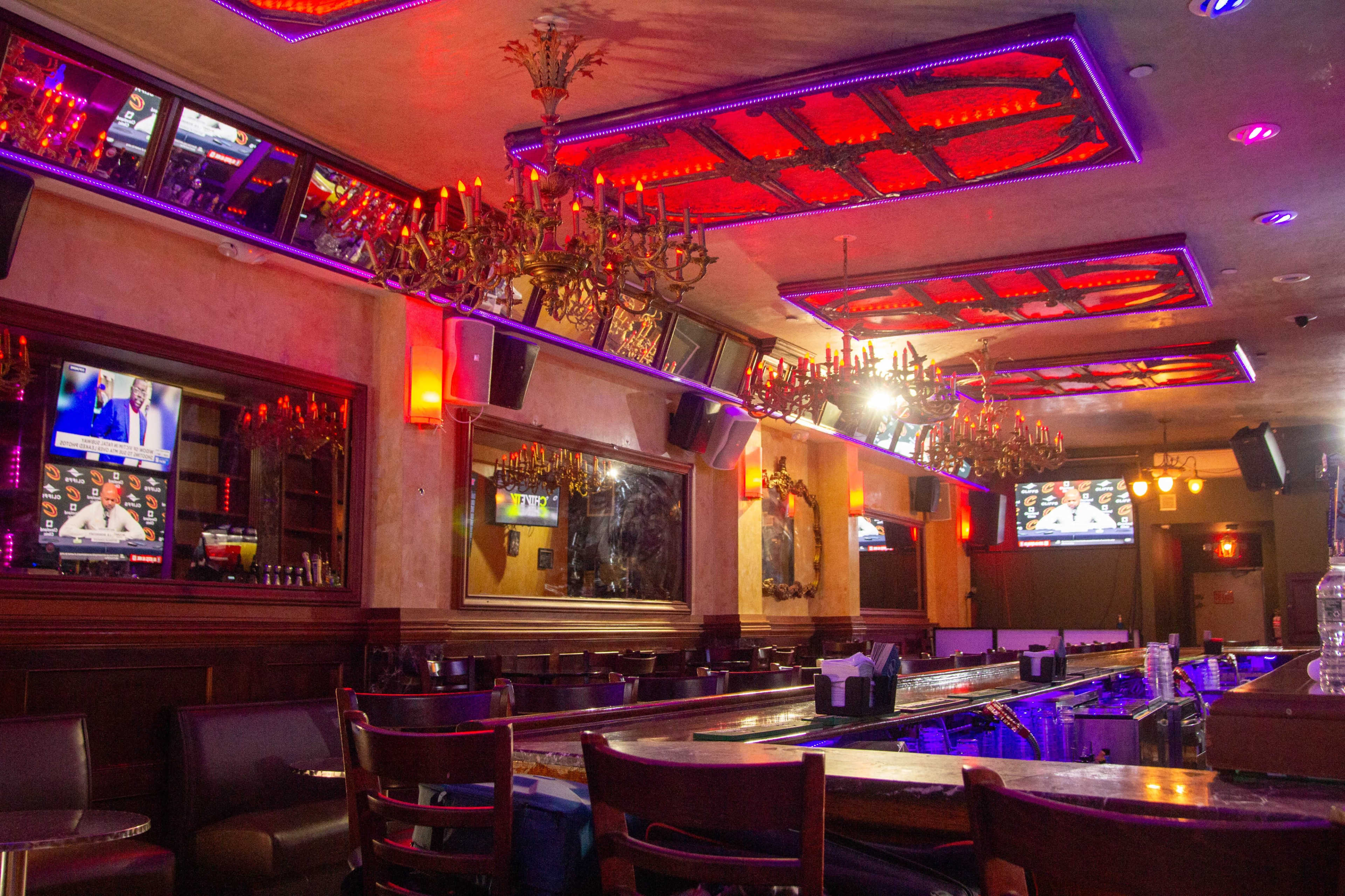 A dimly lit bar with multiple television screens, ornate chandeliers, and a long counter.