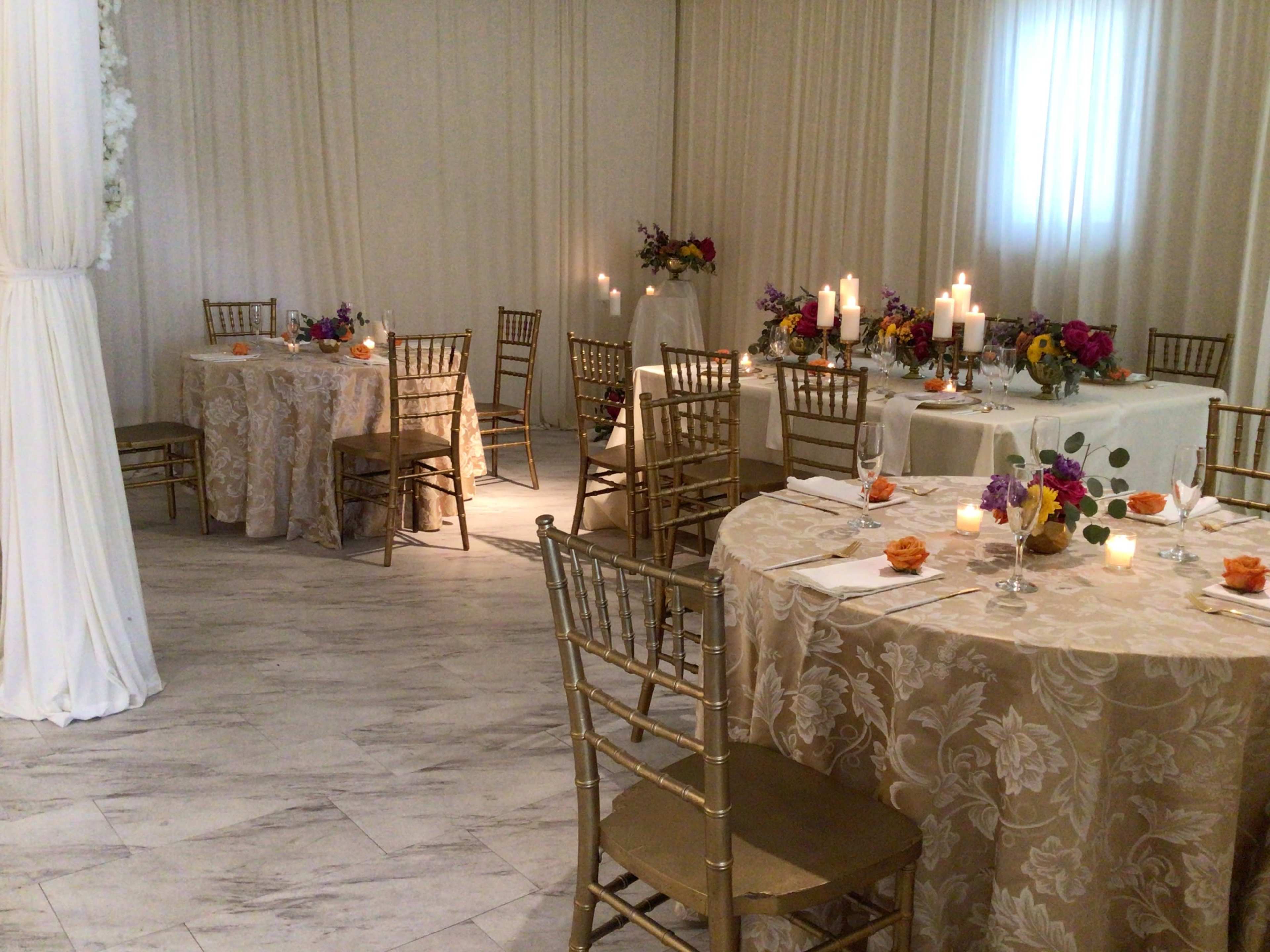 The image shows a decorated event space with several round tables covered in patterned tablecloths, each set with floral centerpieces and candles.