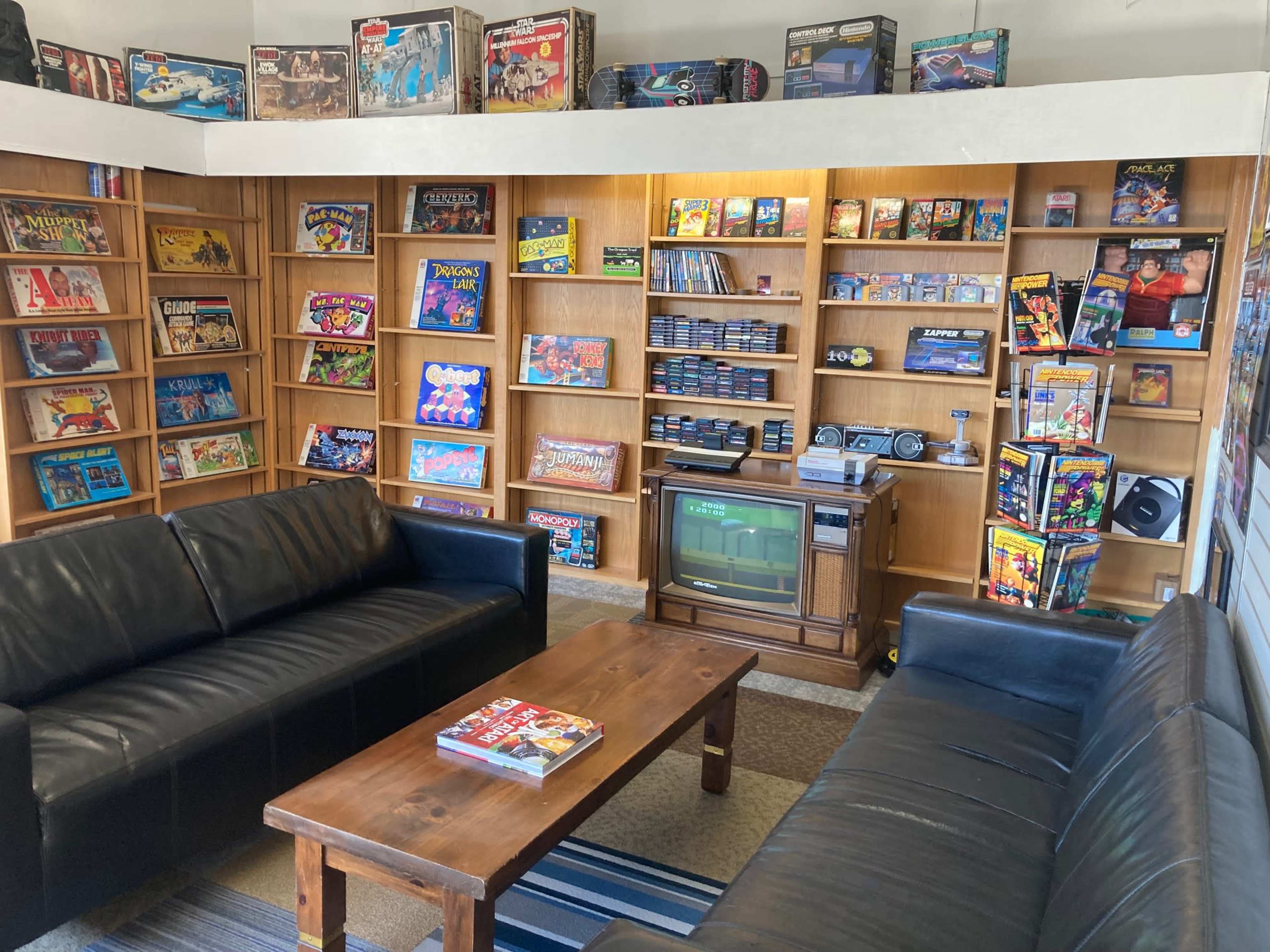 The image shows a cozy game room with two black sofas facing a wooden coffee table, surrounded by shelves filled with board games and video game consoles.