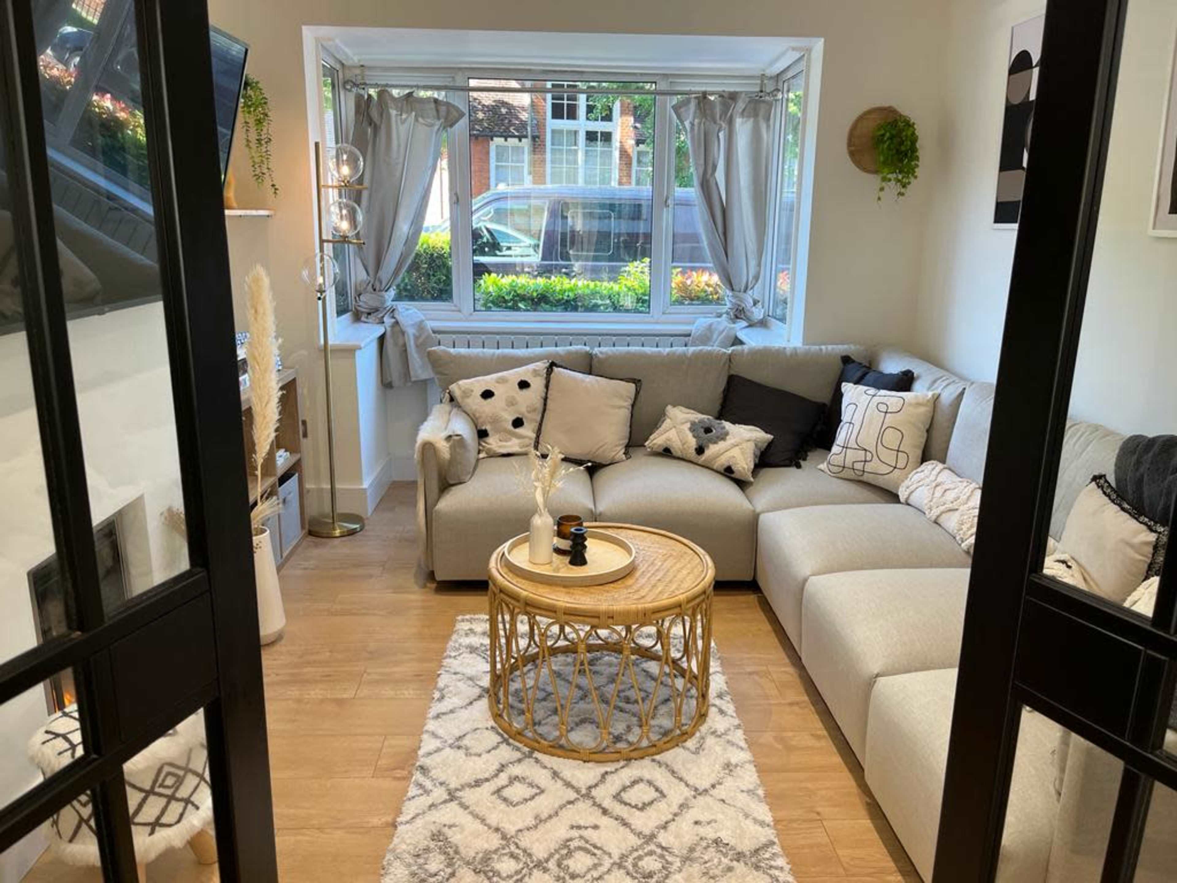 The image shows a modern living room with a sectional sofa, a round coffee table, decorative throw pillows, and large windows overlooking a green park-like area.