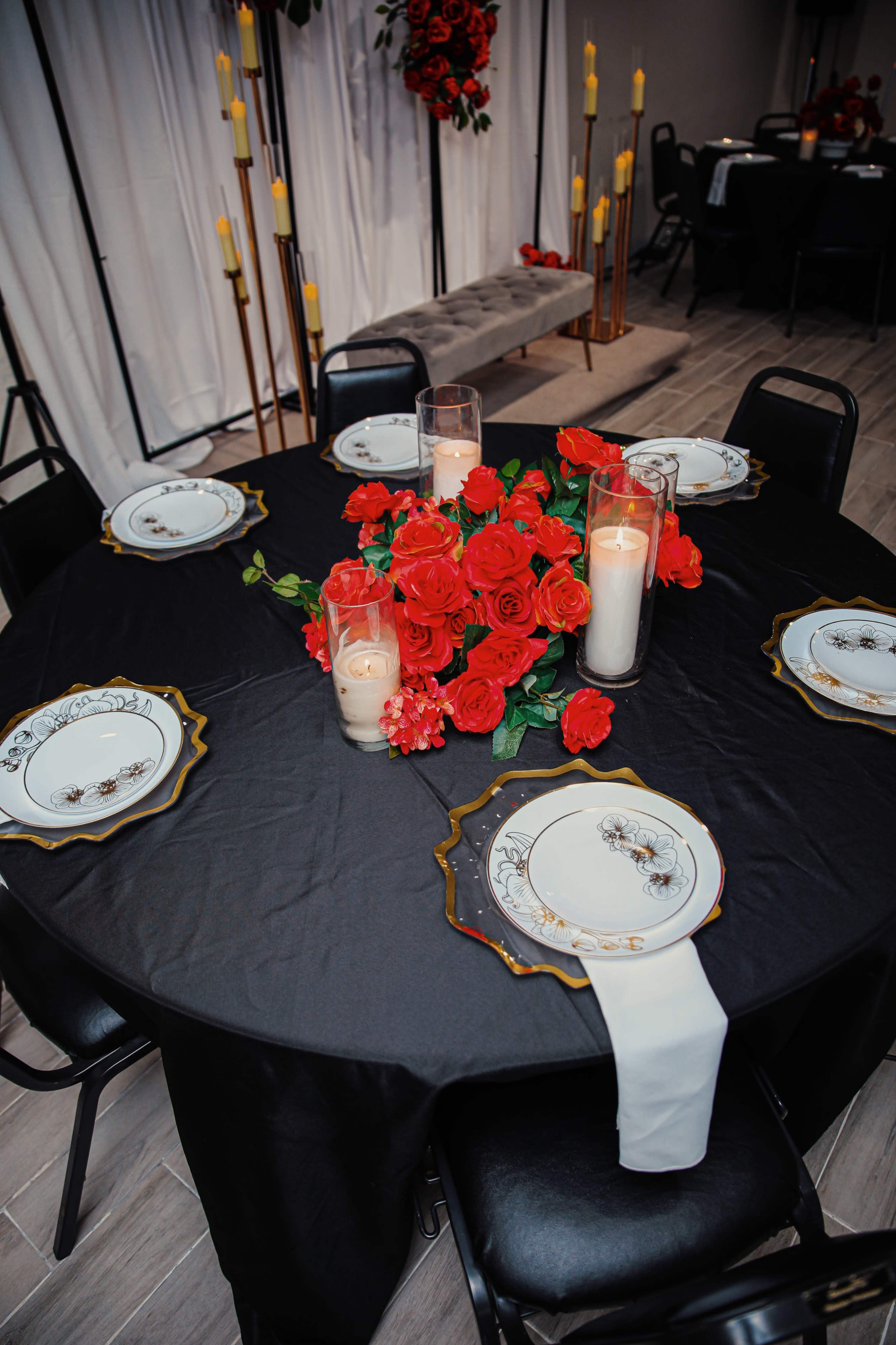 A round black table is set with white plates, red roses, and candles, creating a decorative centerpiece in a dimly lit room.