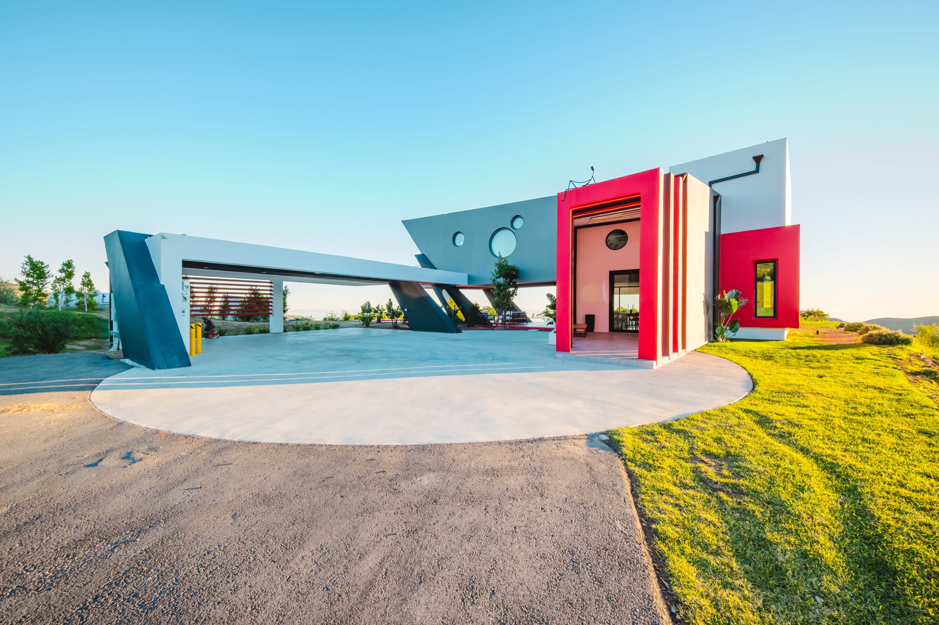 The image shows a modern architectural home with angular shapes, featuring a circular driveway and vibrant red and gray accents.