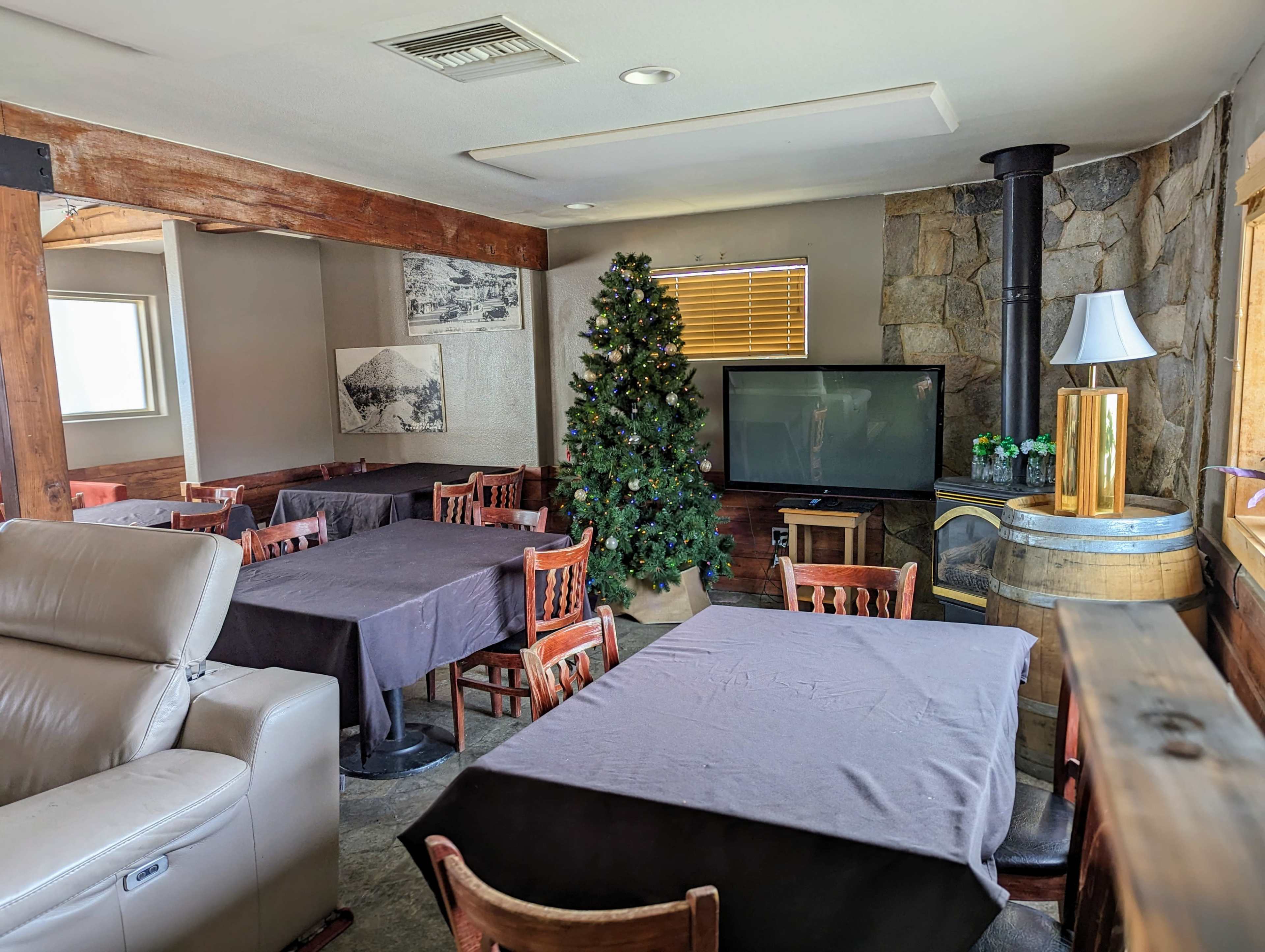 The image shows a spacious room with several wooden tables covered in dark tablecloths, a Christmas tree in the corner, a television, and a stone fireplace.