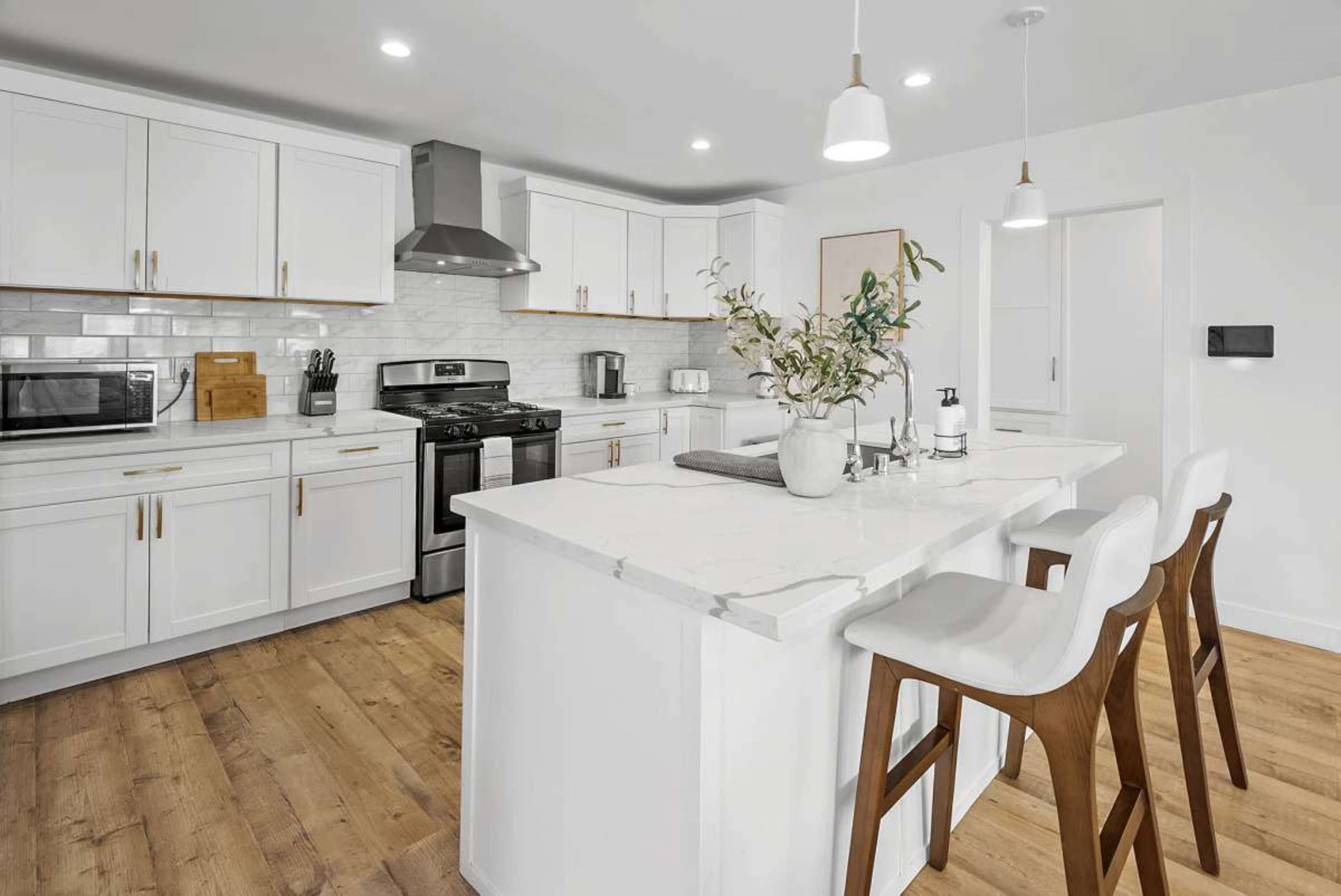 The image shows a modern kitchen featuring white cabinetry, a large island with three barstools, and stainless steel appliances.