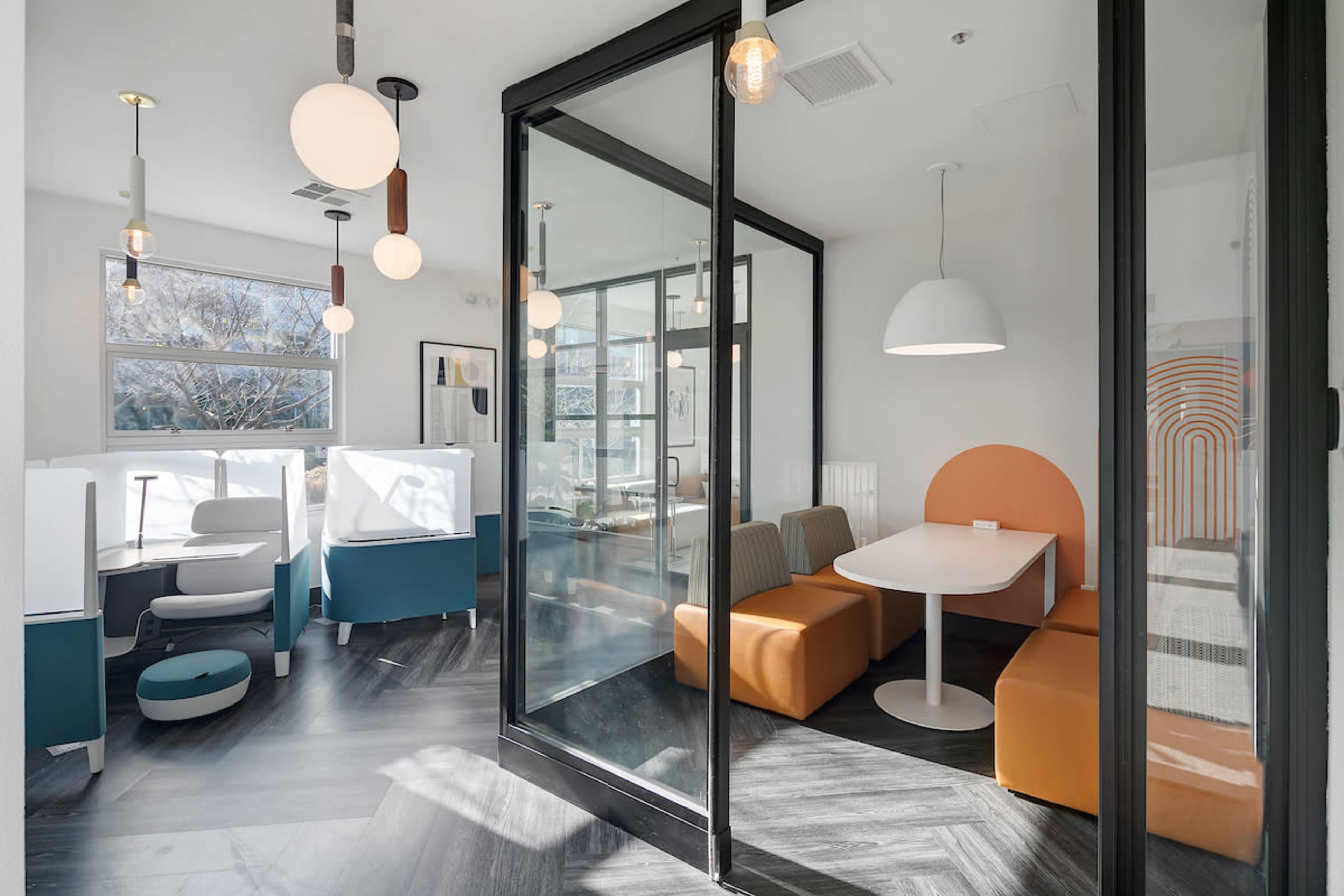 A modern office space featuring a private meeting area with glass walls, a white table, and orange and gray seating.