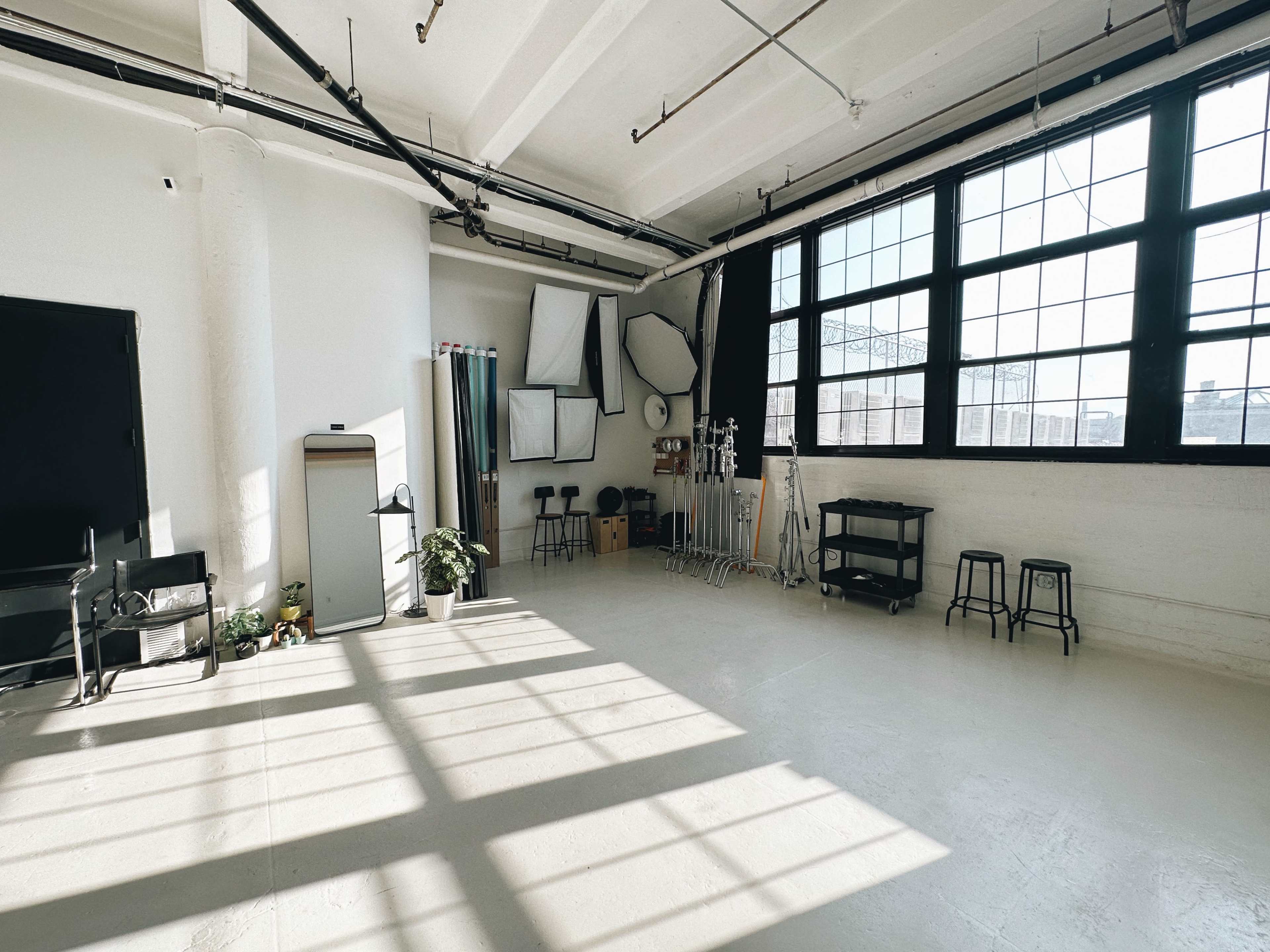 The image shows a spacious, well-lit studio with large windows, reflecting shadows on the floor and featuring various photography equipment and a small plant.