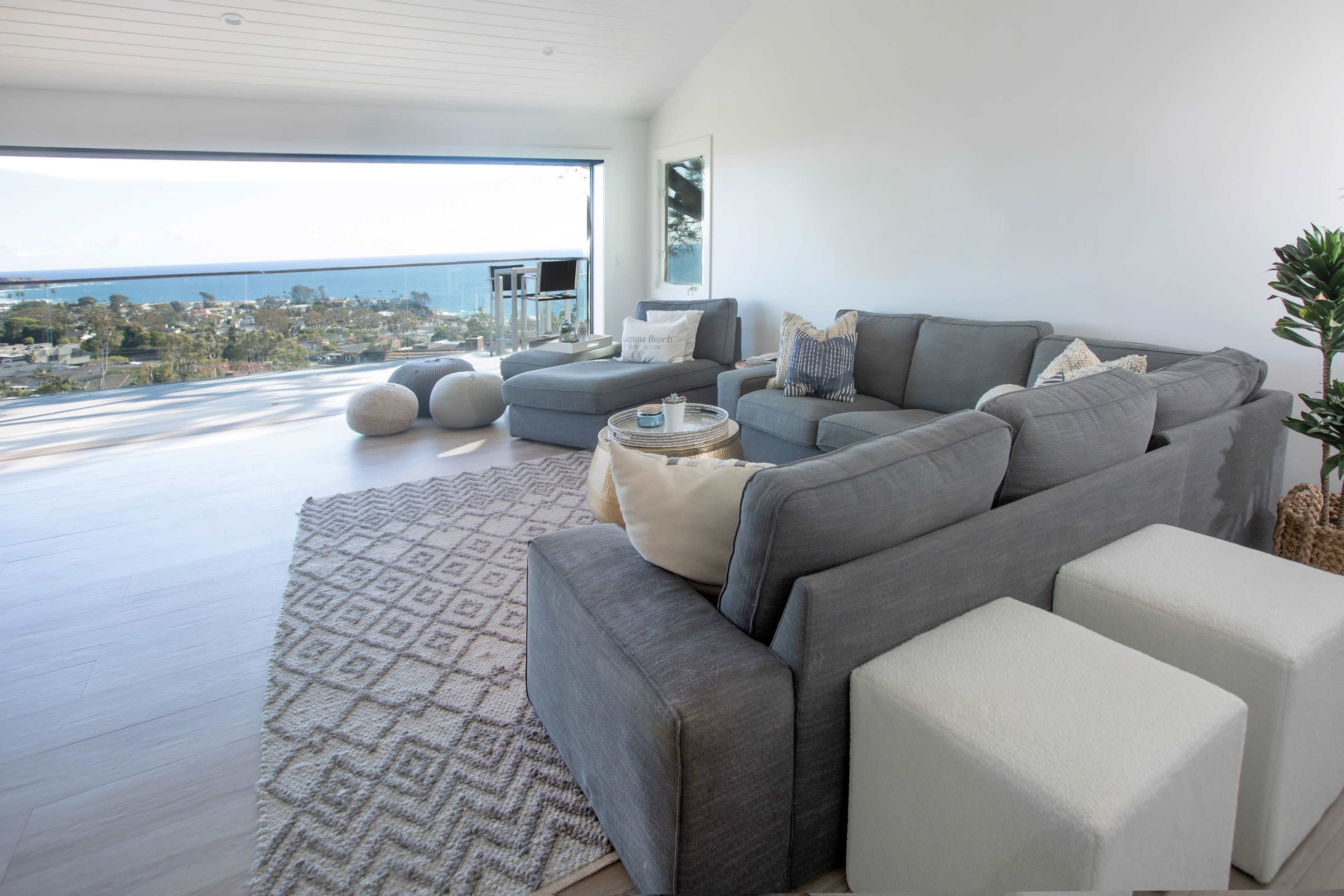 The image shows a modern living room with gray sofas, a large window offering a coastal view, and light-colored decor.