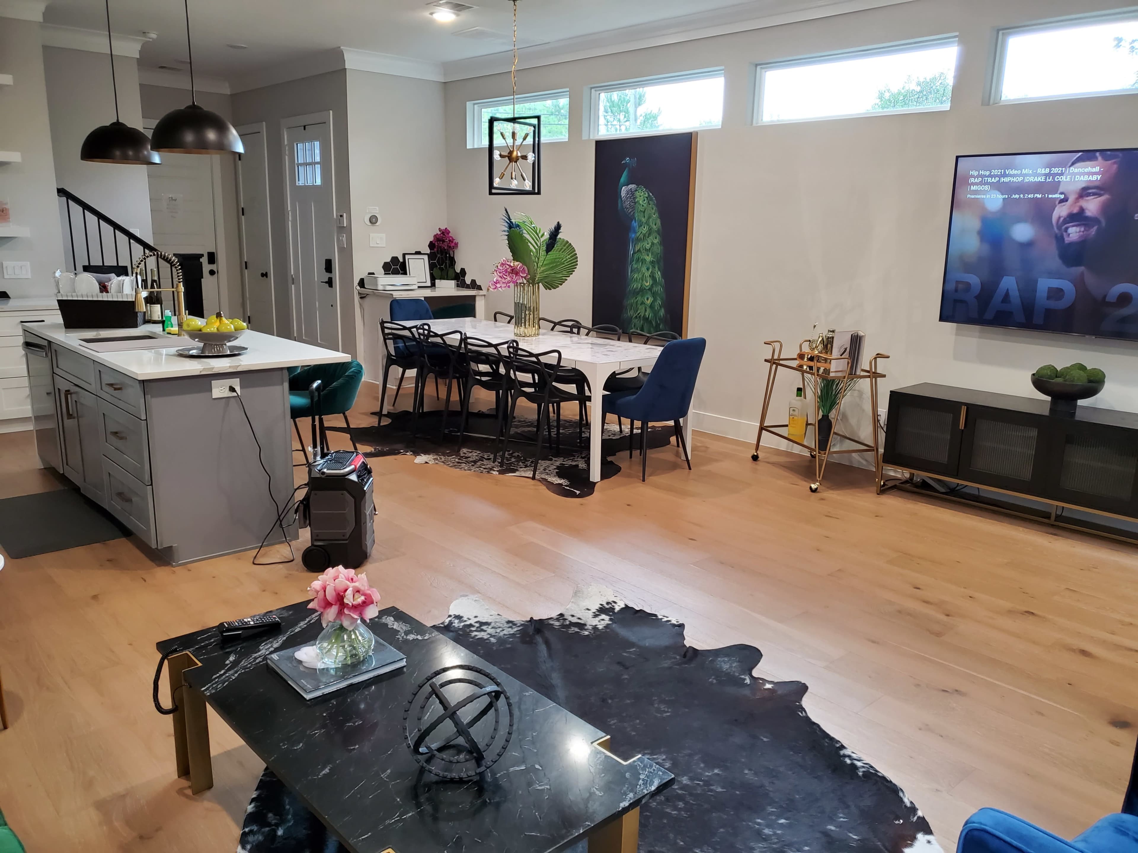 A modern living space featuring a kitchen area, a dining table with chairs, a decorative peacock painting, and a television displaying content on a wooden floor.