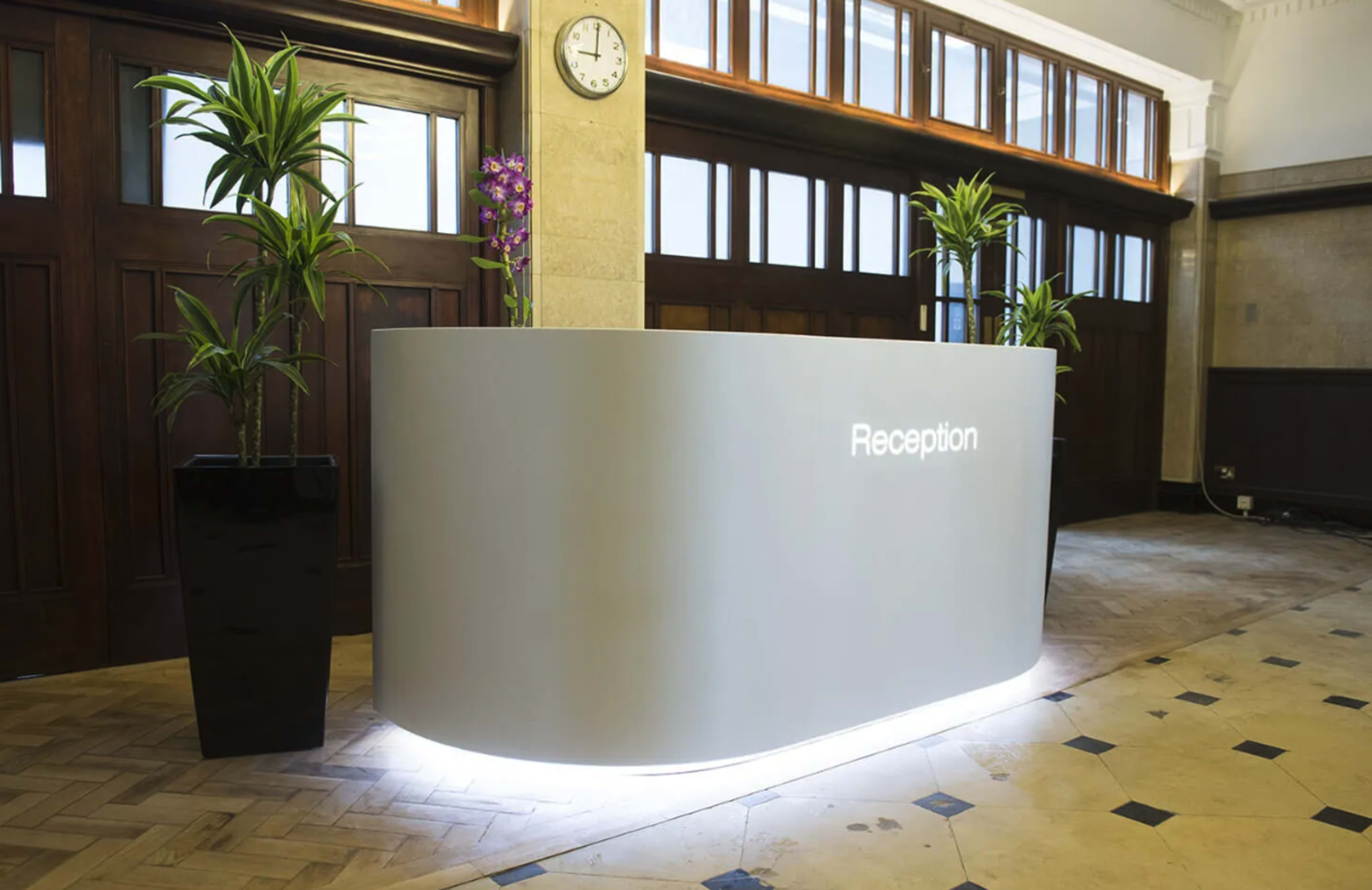 The image shows a modern reception desk with the word "Reception" illuminated, surrounded by potted plants, in a well-lit lobby area.