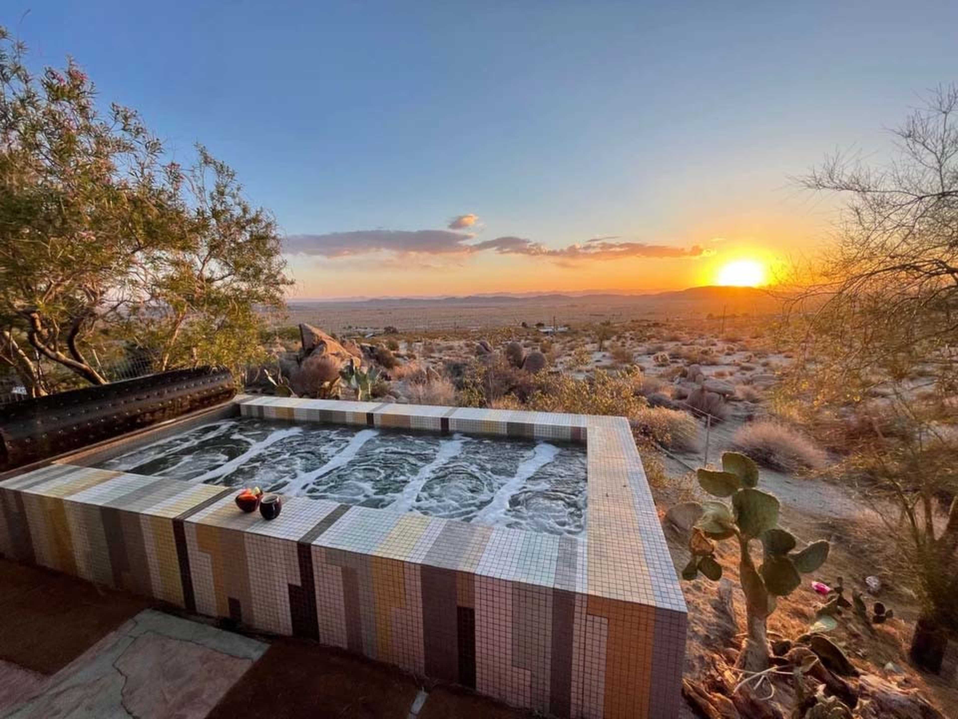 A hot tub overlooks a desert landscape under a setting sun, with cacti and rocks surrounding the area.