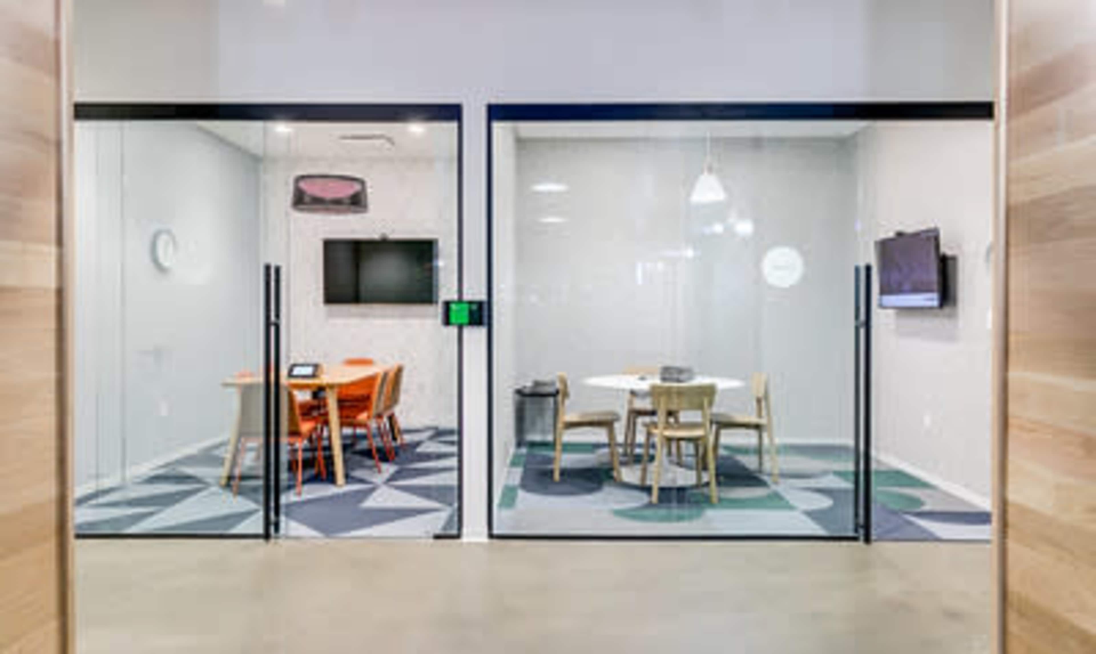 The image shows two glass-walled meeting rooms, one with a table and orange chairs and the other with a round table and wooden chairs.