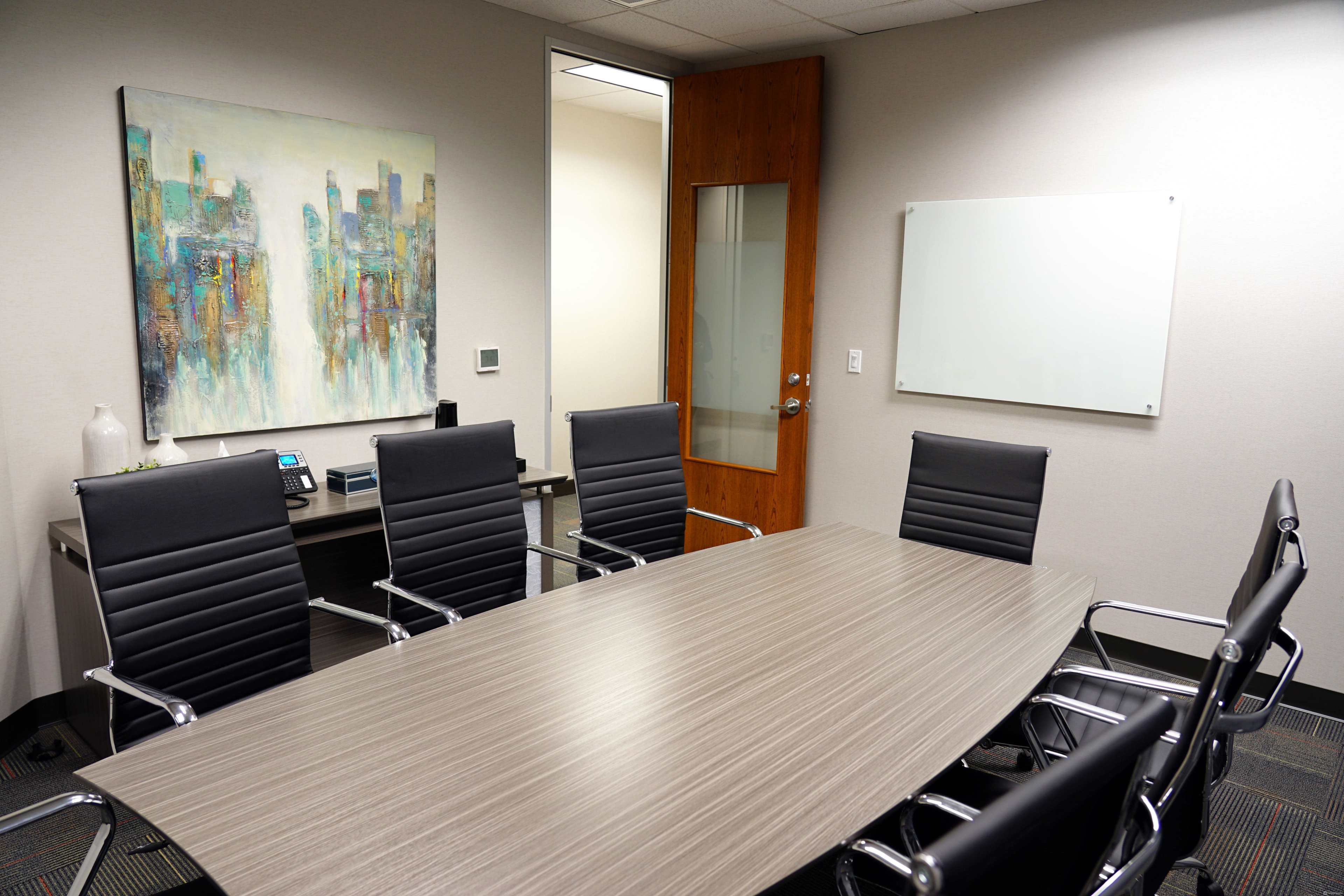 A modern conference room with a large table surrounded by black chairs, a painting on the wall, and a whiteboard next to a glass door.