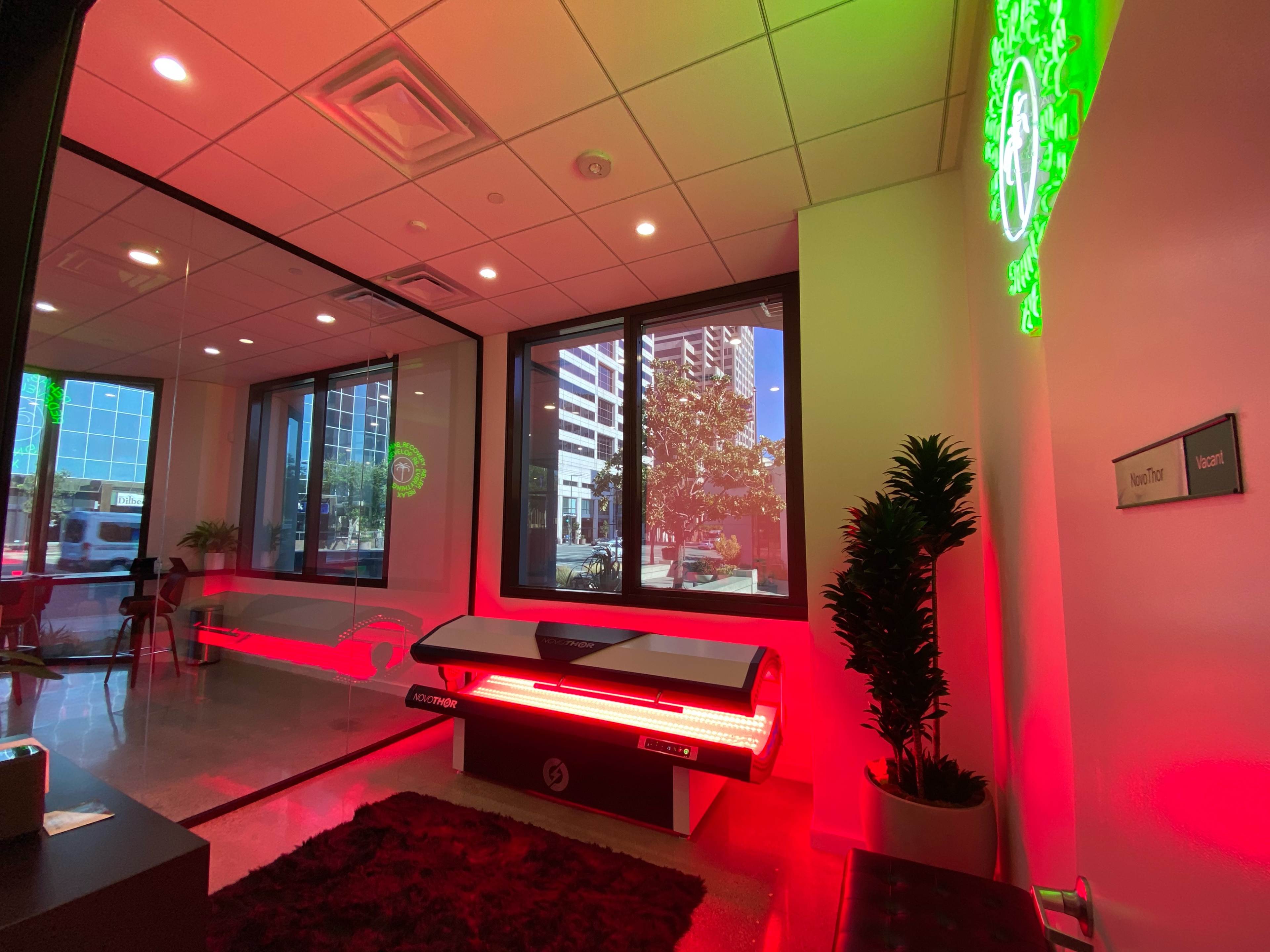 The image shows a modern wellness facility with a red light therapy bed positioned near large windows overlooking an urban landscape.