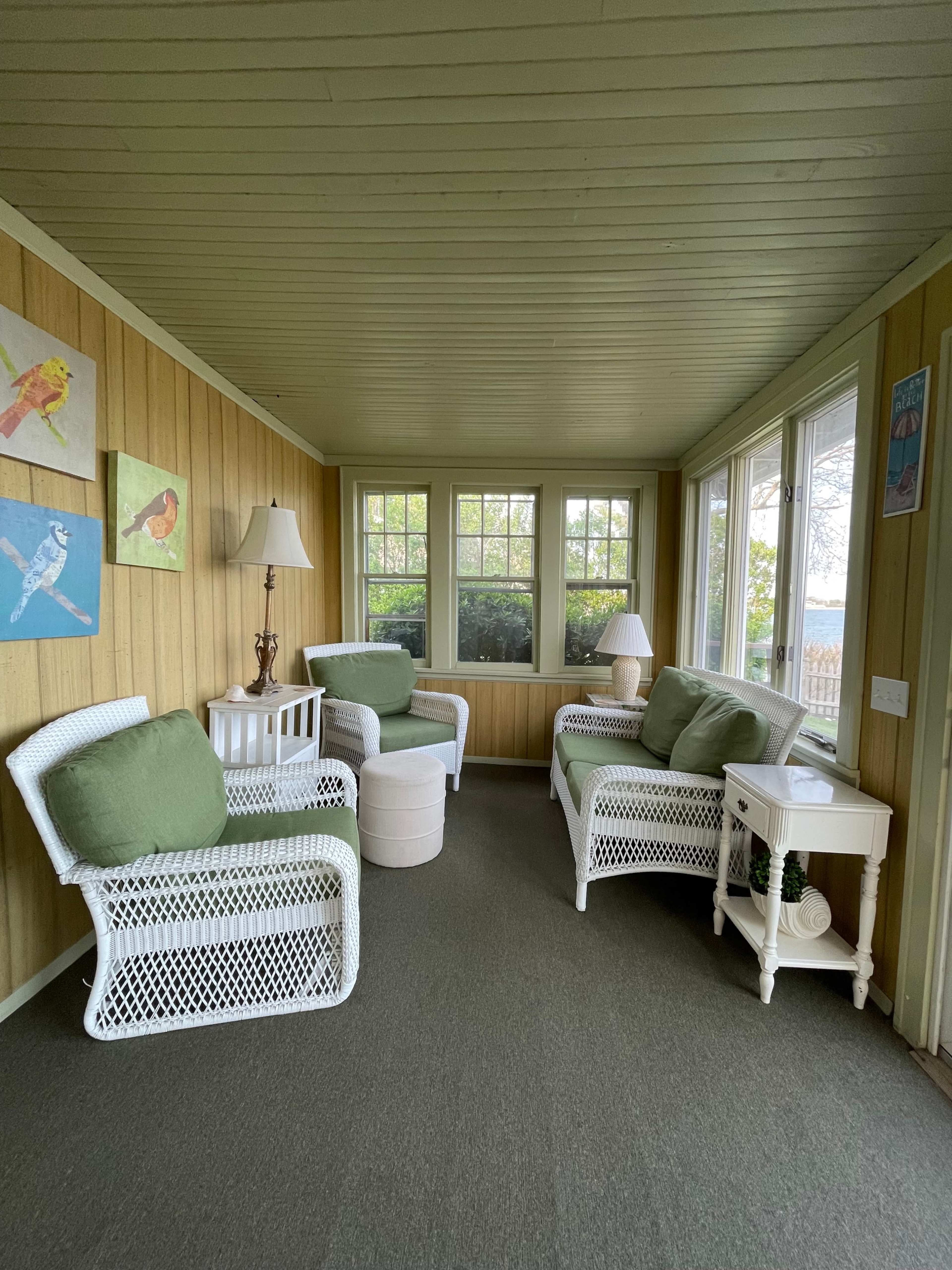 A sunroom with white wicker furniture, green cushions, and wooden walls, featuring windows that overlook an outdoor view.
