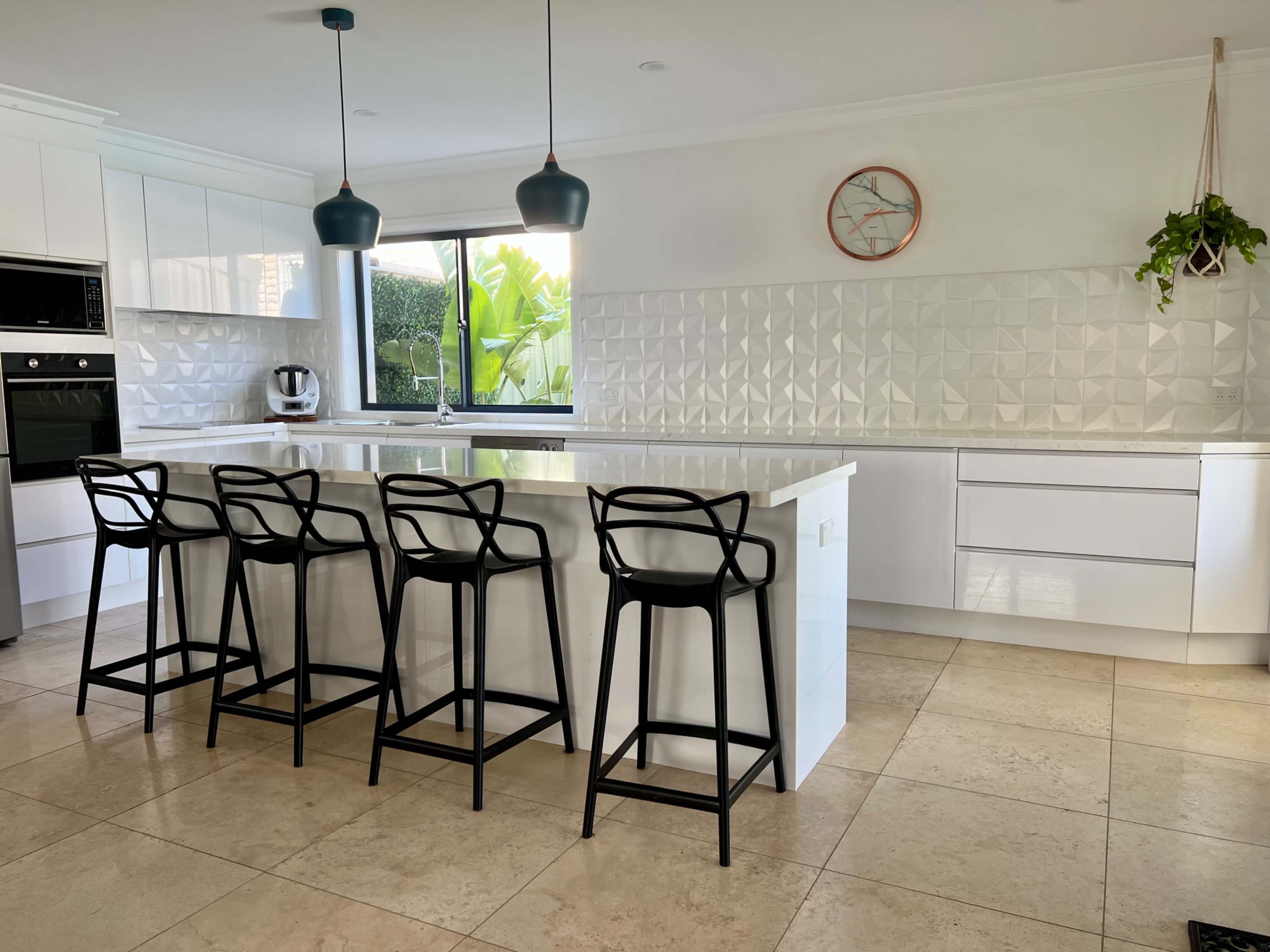 A modern kitchen features a large island with four black bar stools, white cabinetry, and geometric backsplash tiles.
