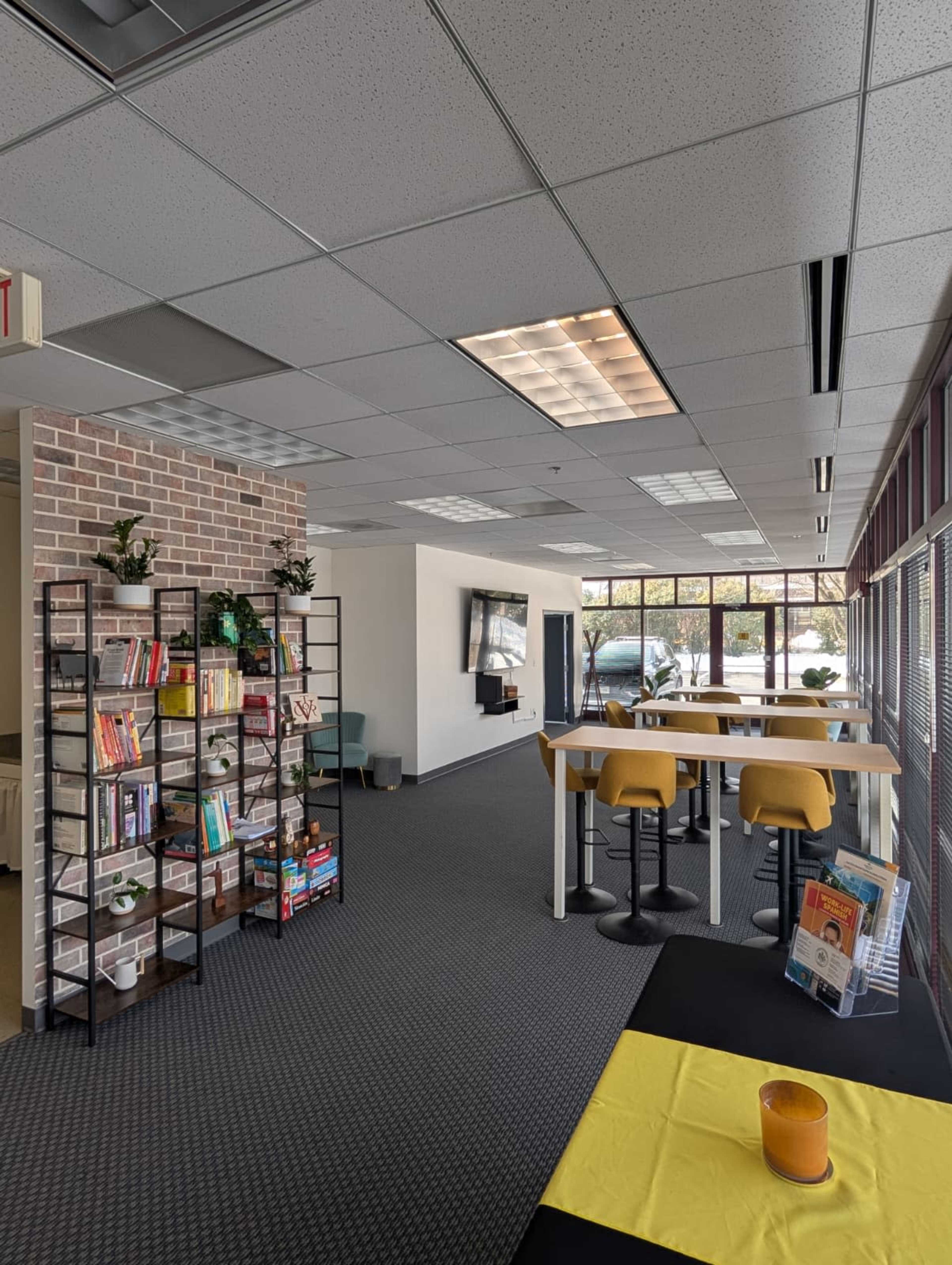The image shows a modern office space featuring a bookshelf filled with various items, a bar-height table with yellow stools, and large windows allowing natural light.