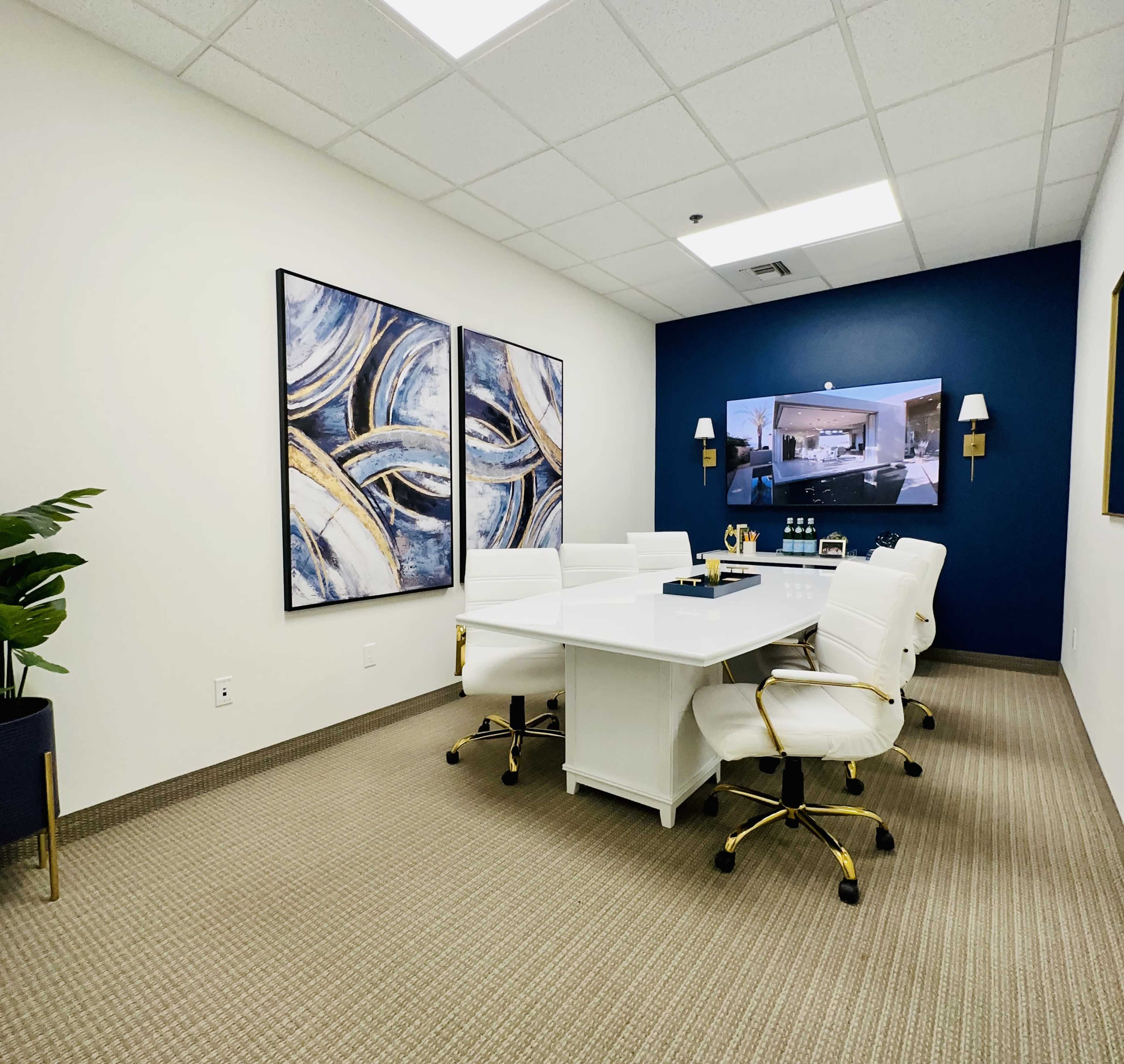 A modern conference room with a white table surrounded by white chairs, complemented by blue wall decor and a flat-screen monitor.