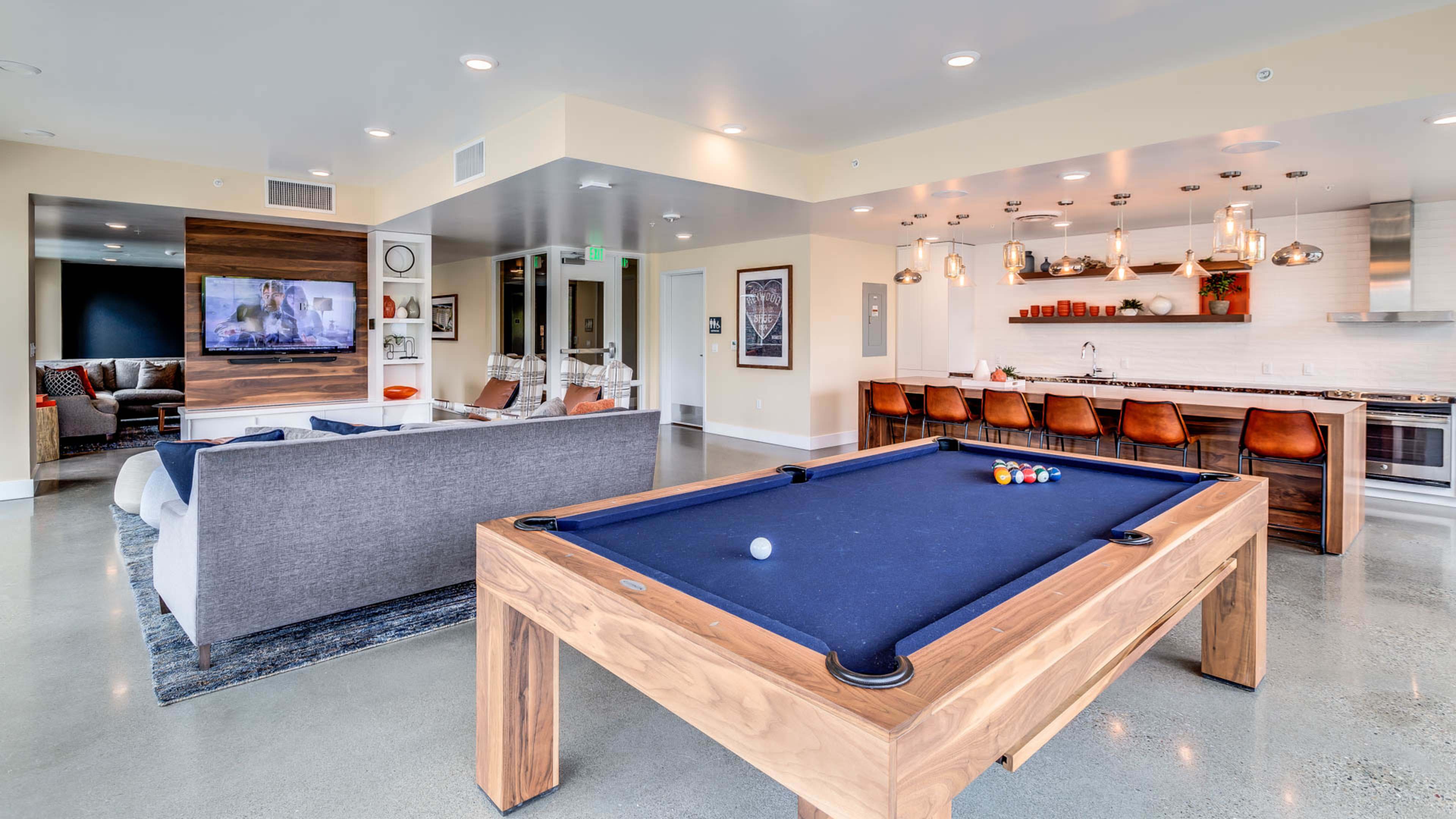 The image shows a modern lounge area featuring a pool table, a seating arrangement, and a kitchen bar with stools.