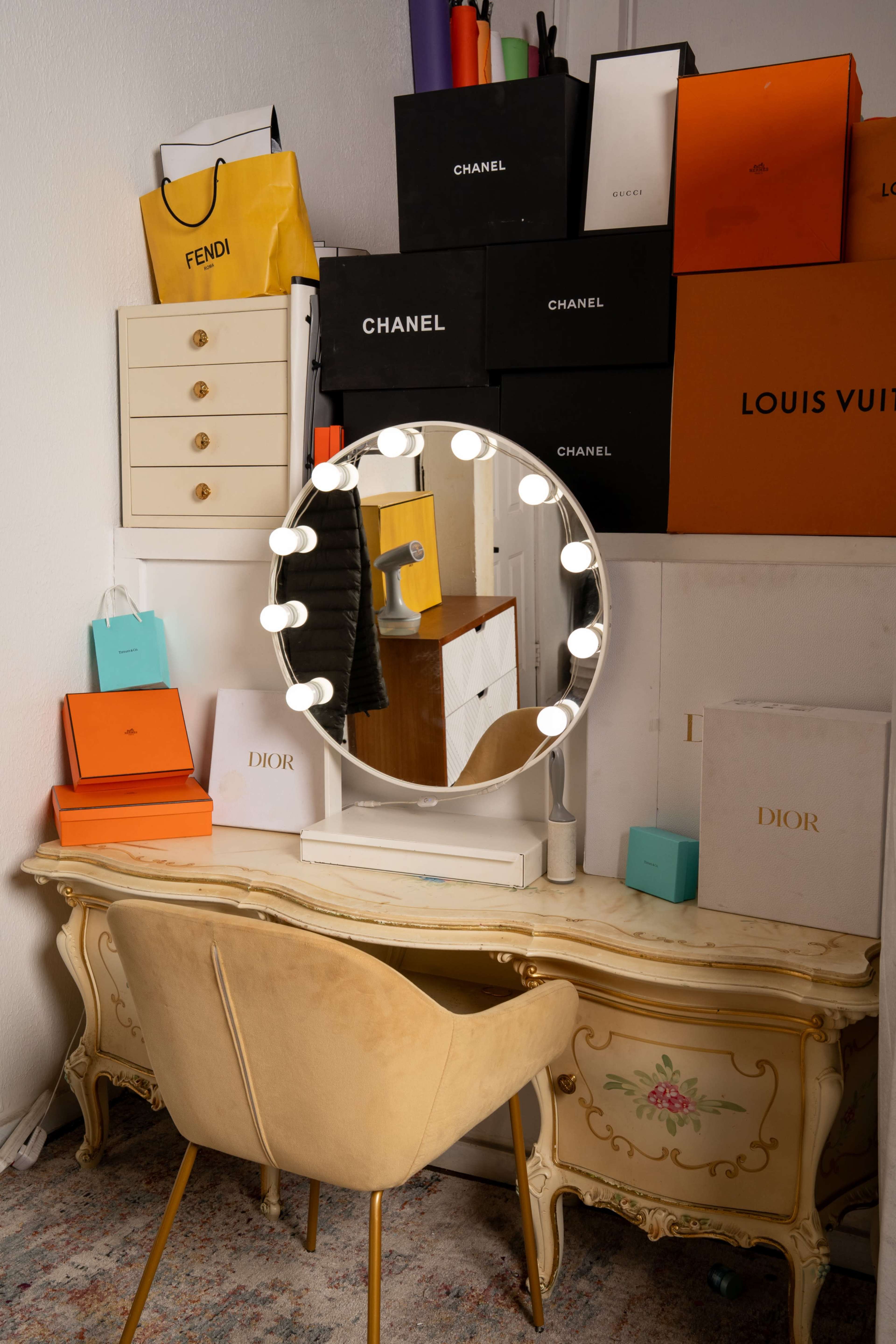 A vanity table with a circular mirror surrounded by various luxury brand boxes and a plush chair sits in a small room.