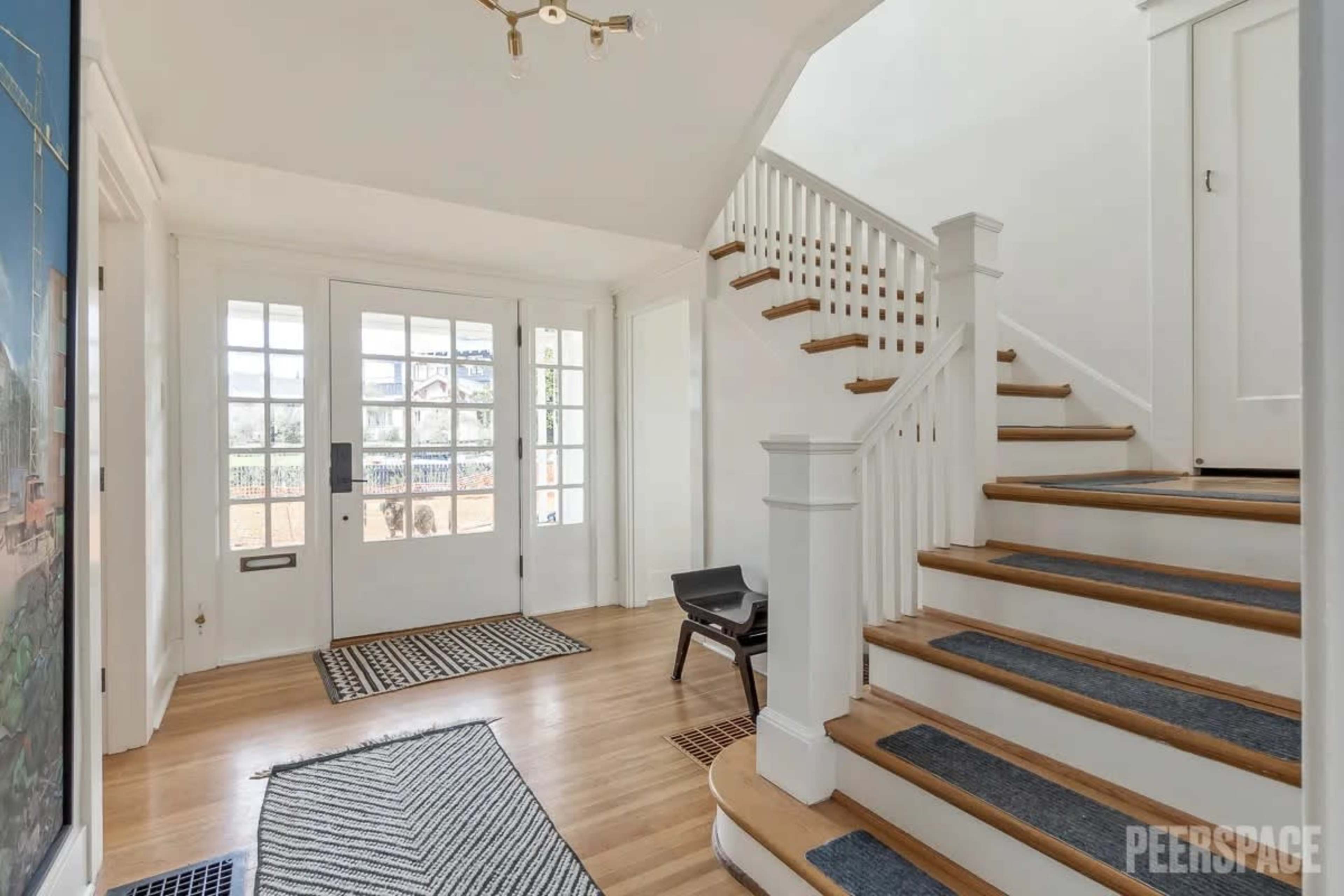 A bright entryway with a staircase on the right, a patterned rug on the floor, and large windows on either side of the front door.