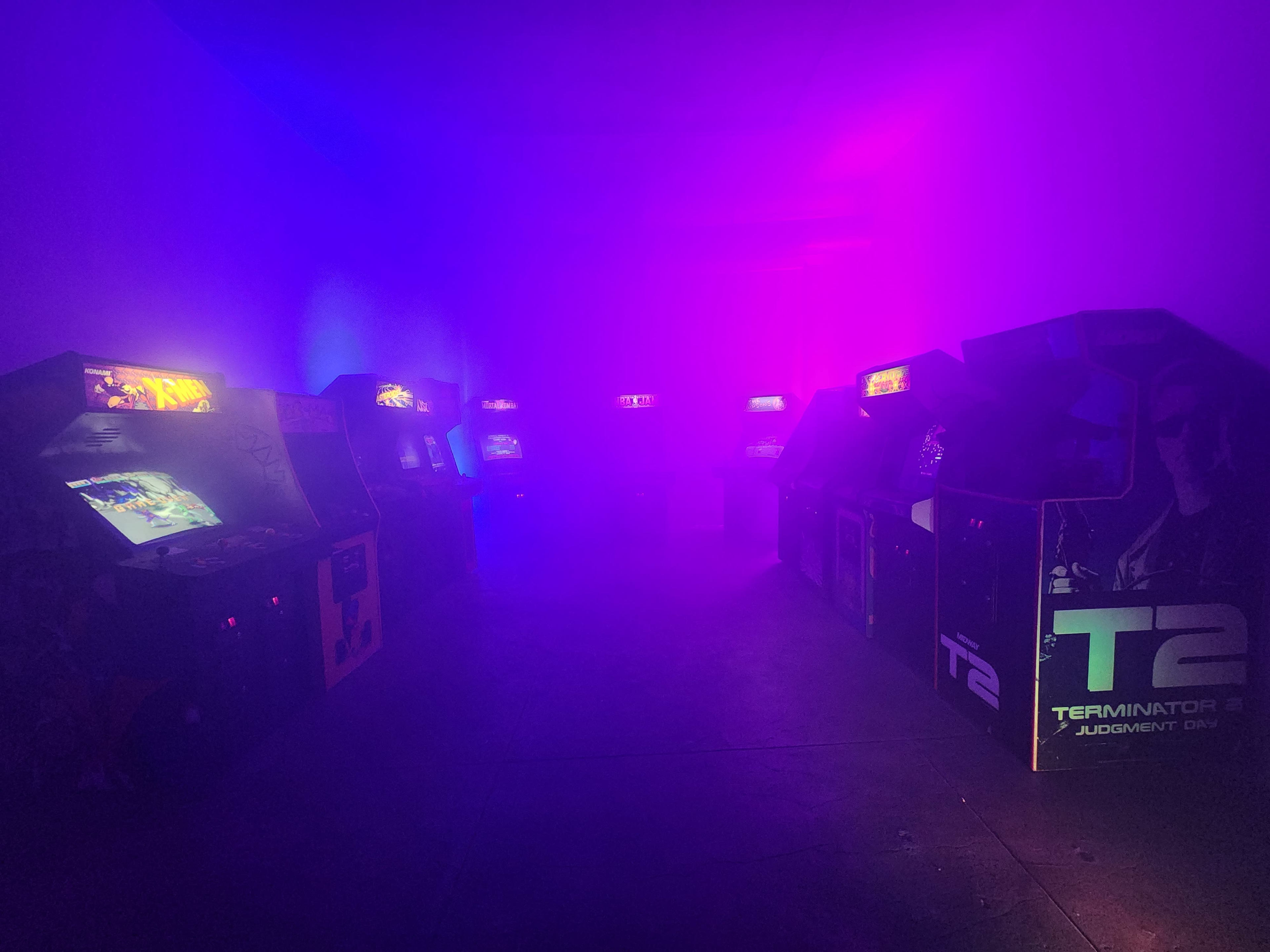 A dimly lit arcade filled with retro arcade machines, illuminated by purple and blue lighting amidst a haze.