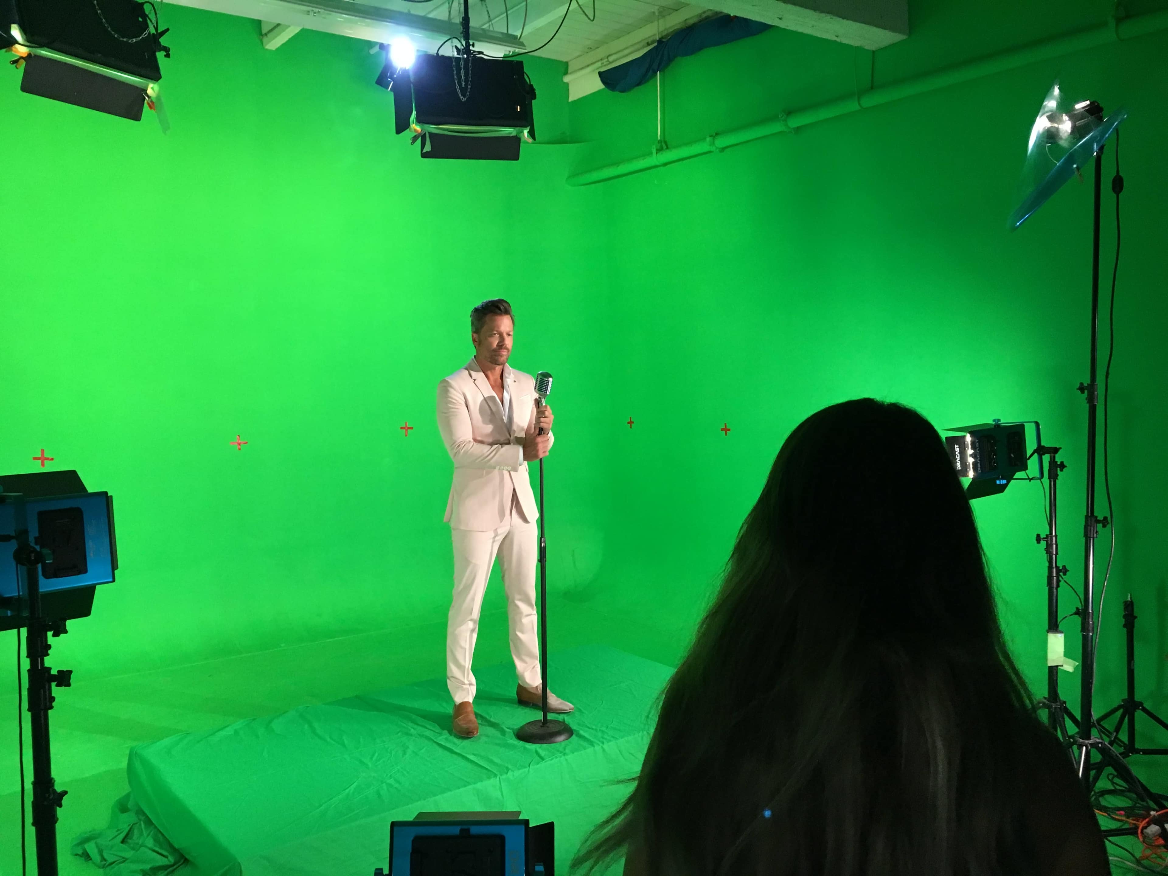 A man in a light-colored suit stands on a green background, holding a microphone, while a woman with long hair observes from behind a camera setup.