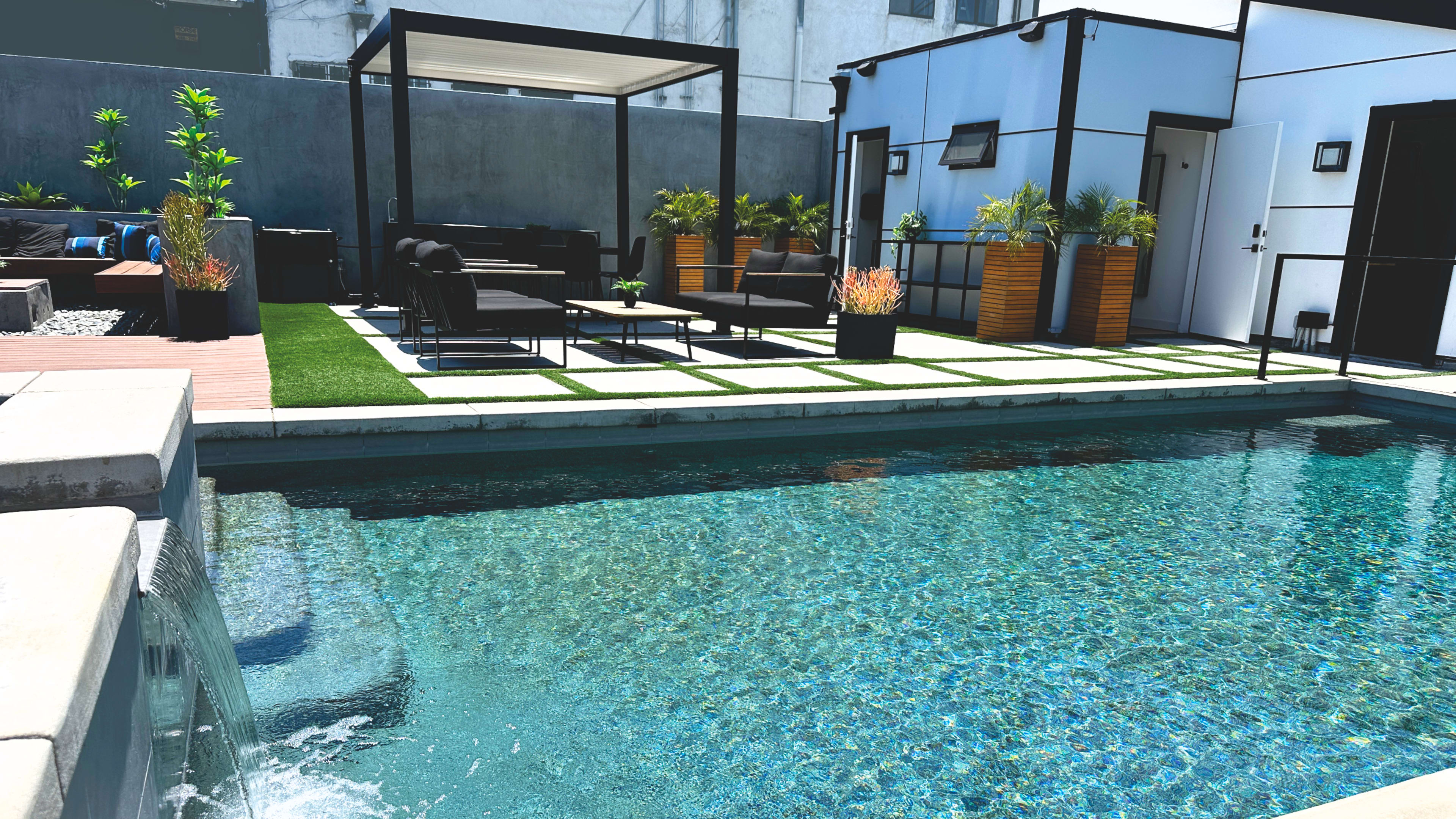 A modern outdoor space featuring a swimming pool, seating areas with black furniture, and lush greenery in planters.