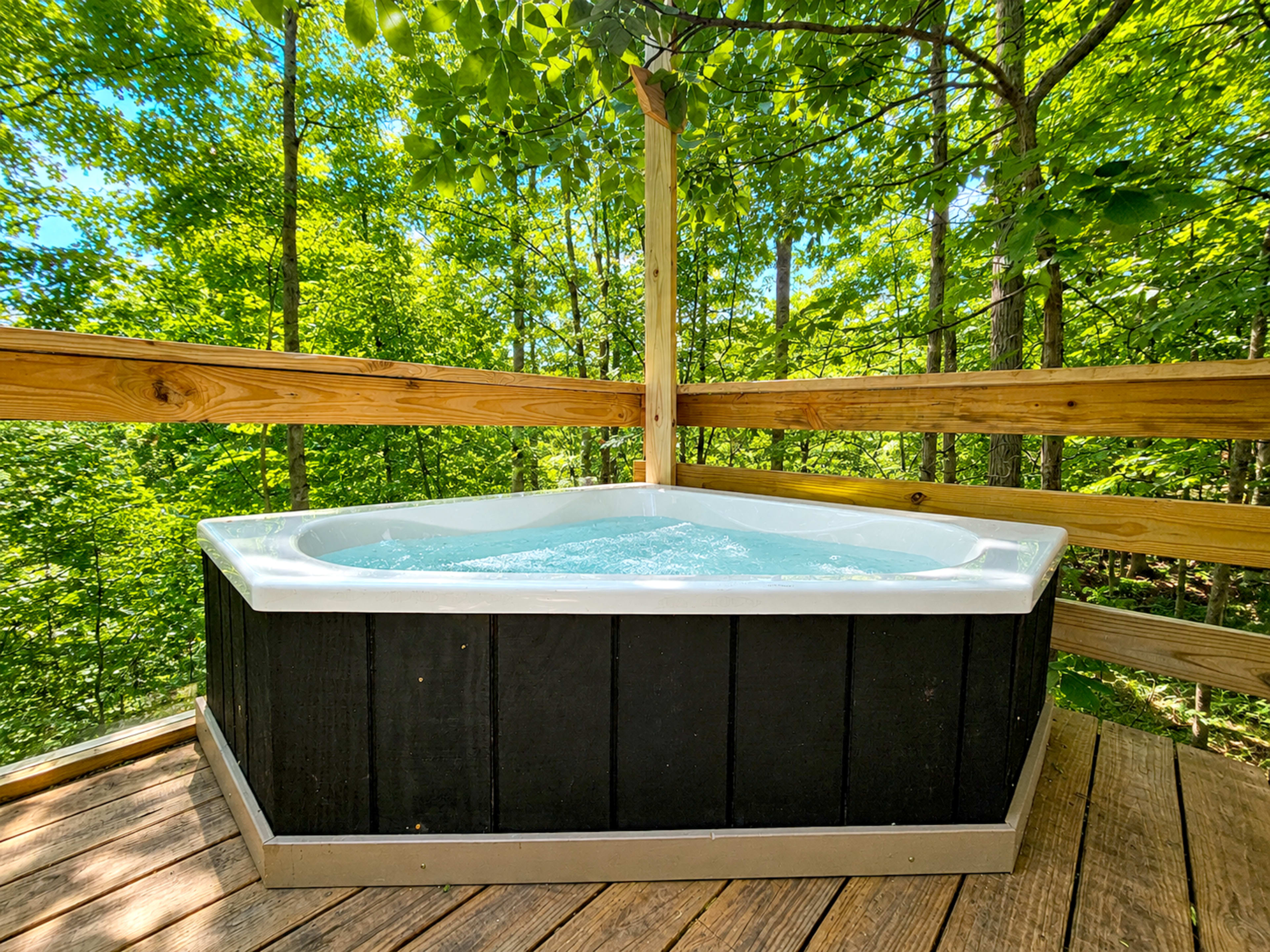 A triangular hot tub filled with water is situated on a wooden deck surrounded by lush green trees.