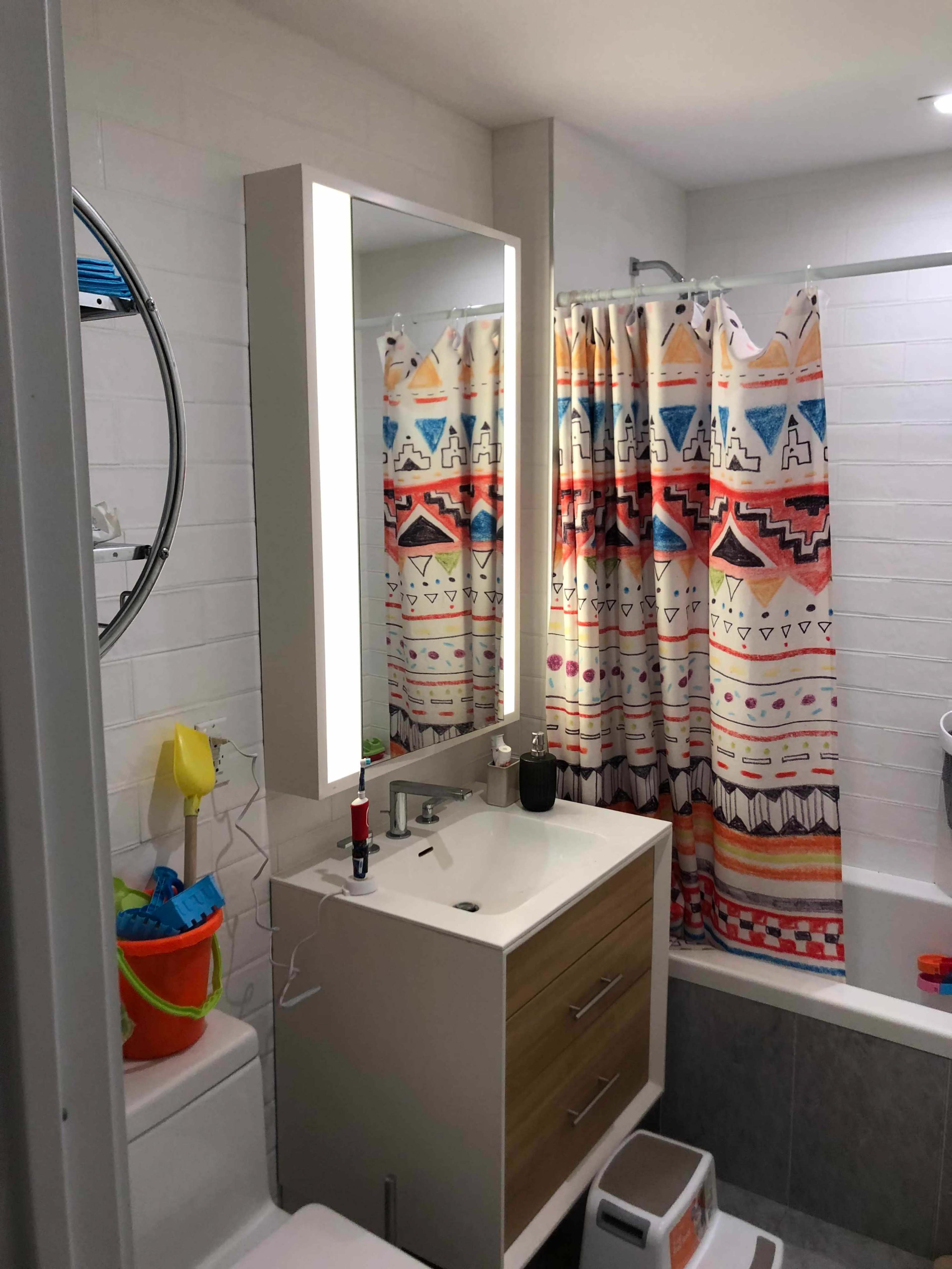 A bathroom features a modern vanity with a lit mirror, colorful shower curtain, and a bathtub with a showerhead.