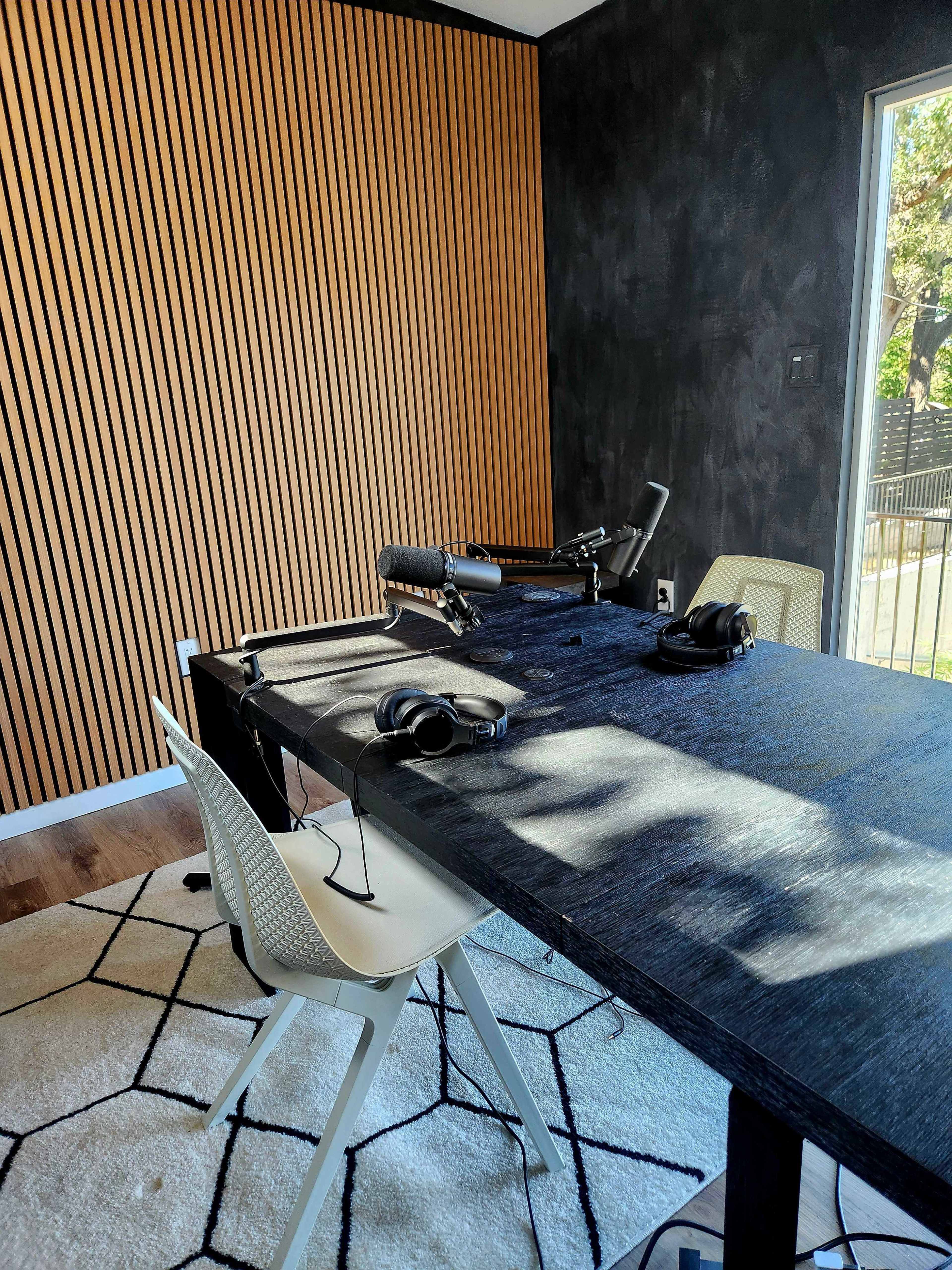 The image shows a modern workspace featuring a desk with headphones placed on it, surrounded by minimalist white chairs and a wood-paneled accent wall.