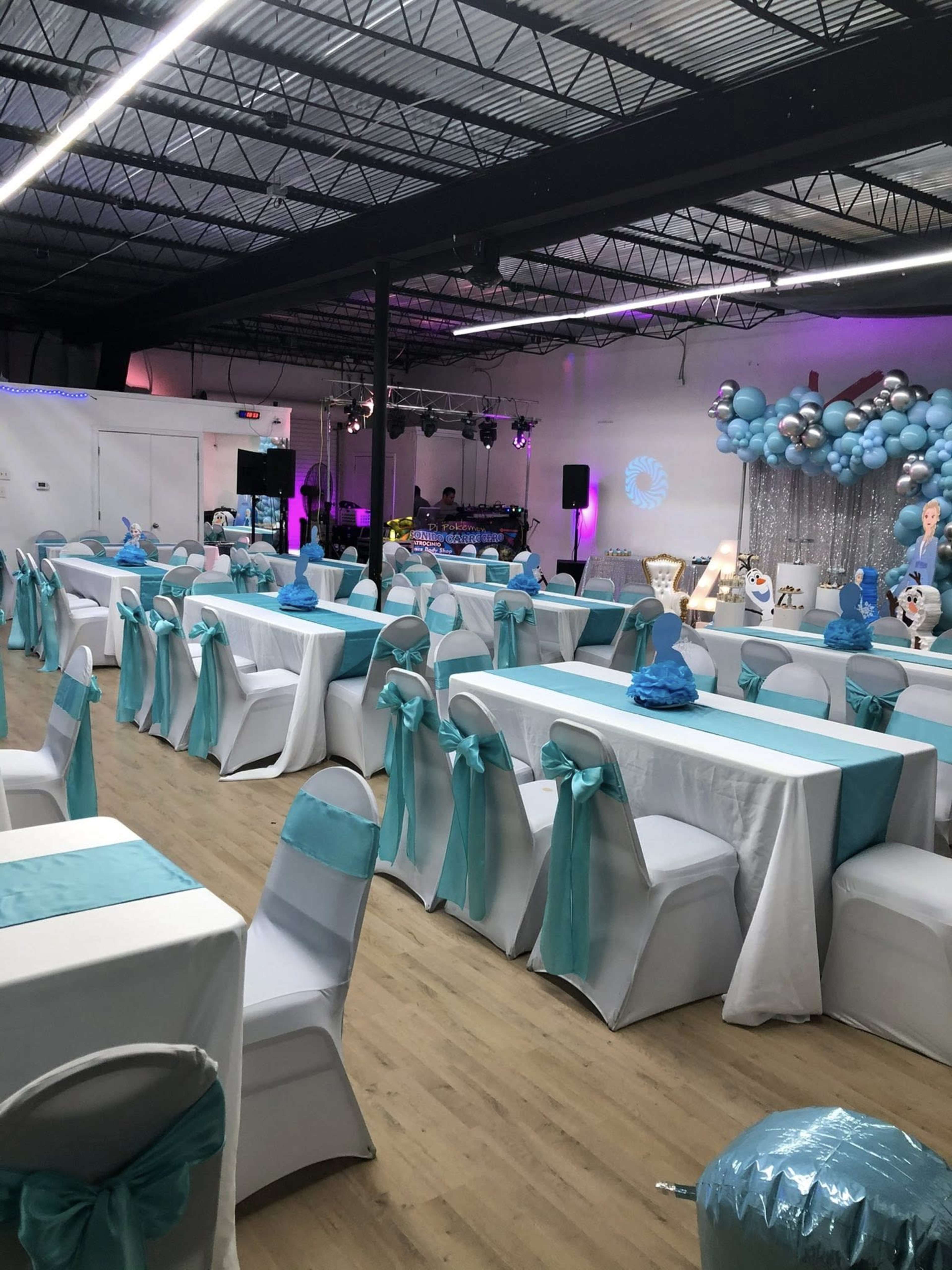 The image shows a decorated event space with tables covered in white linens and turquoise accents, set up for a celebration with balloons and a stage in the background.