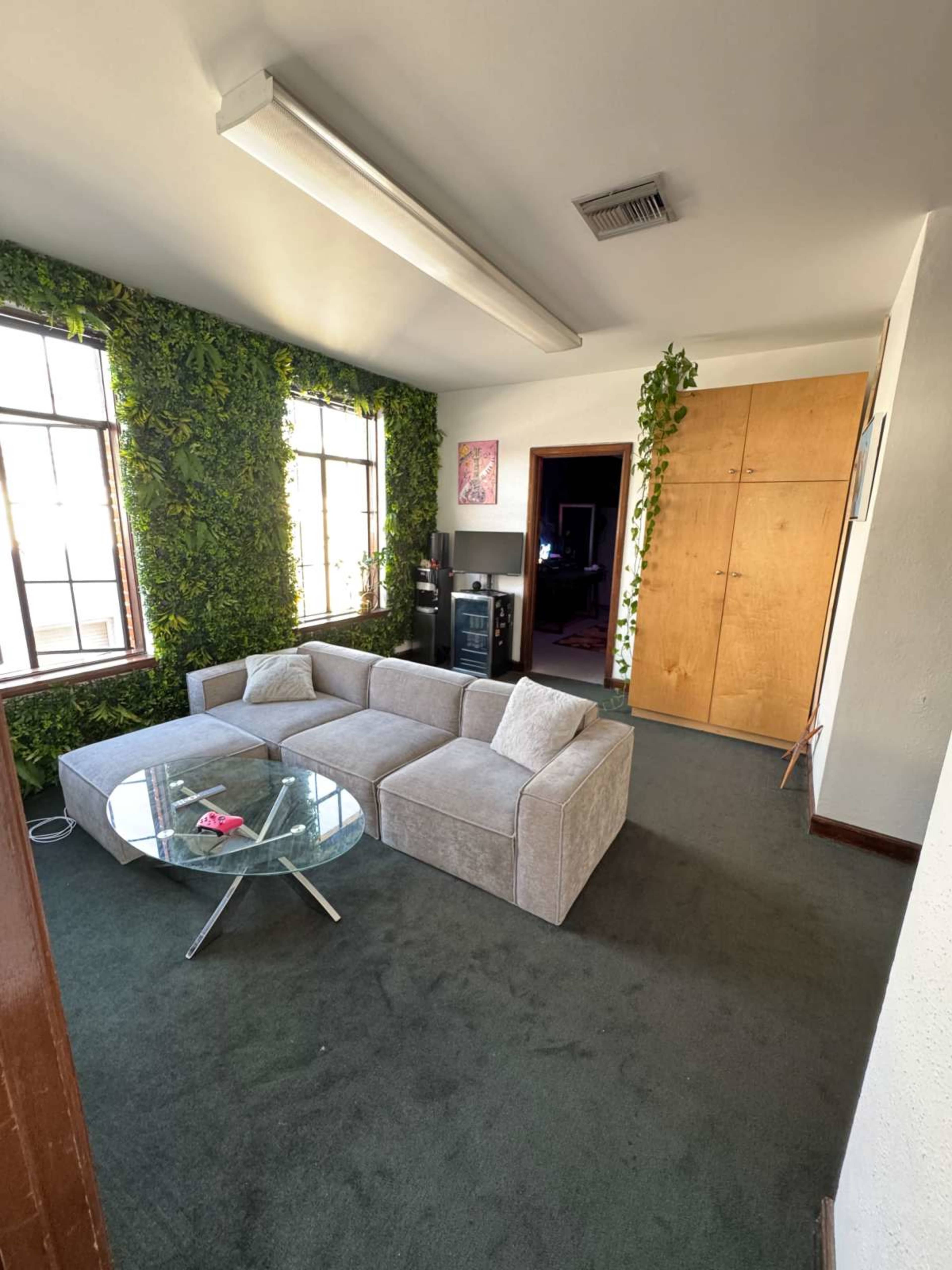 The image shows a living room with a gray sectional sofa, a glass coffee table, and green plants adorning the walls.