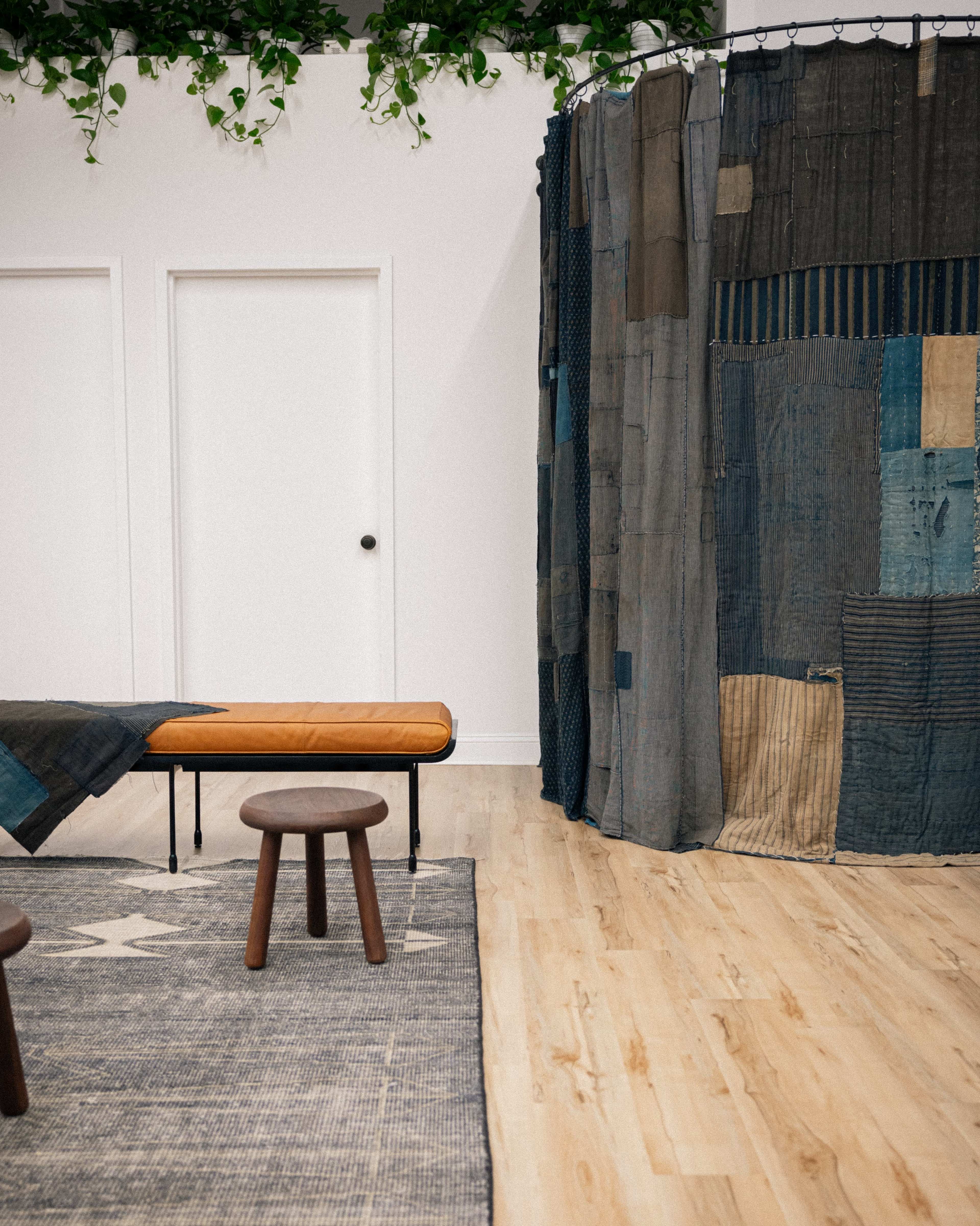 The image shows a modern room featuring a wooden floor, a bench with a leather seat, a round stool, and a curtain made of patchwork fabric.