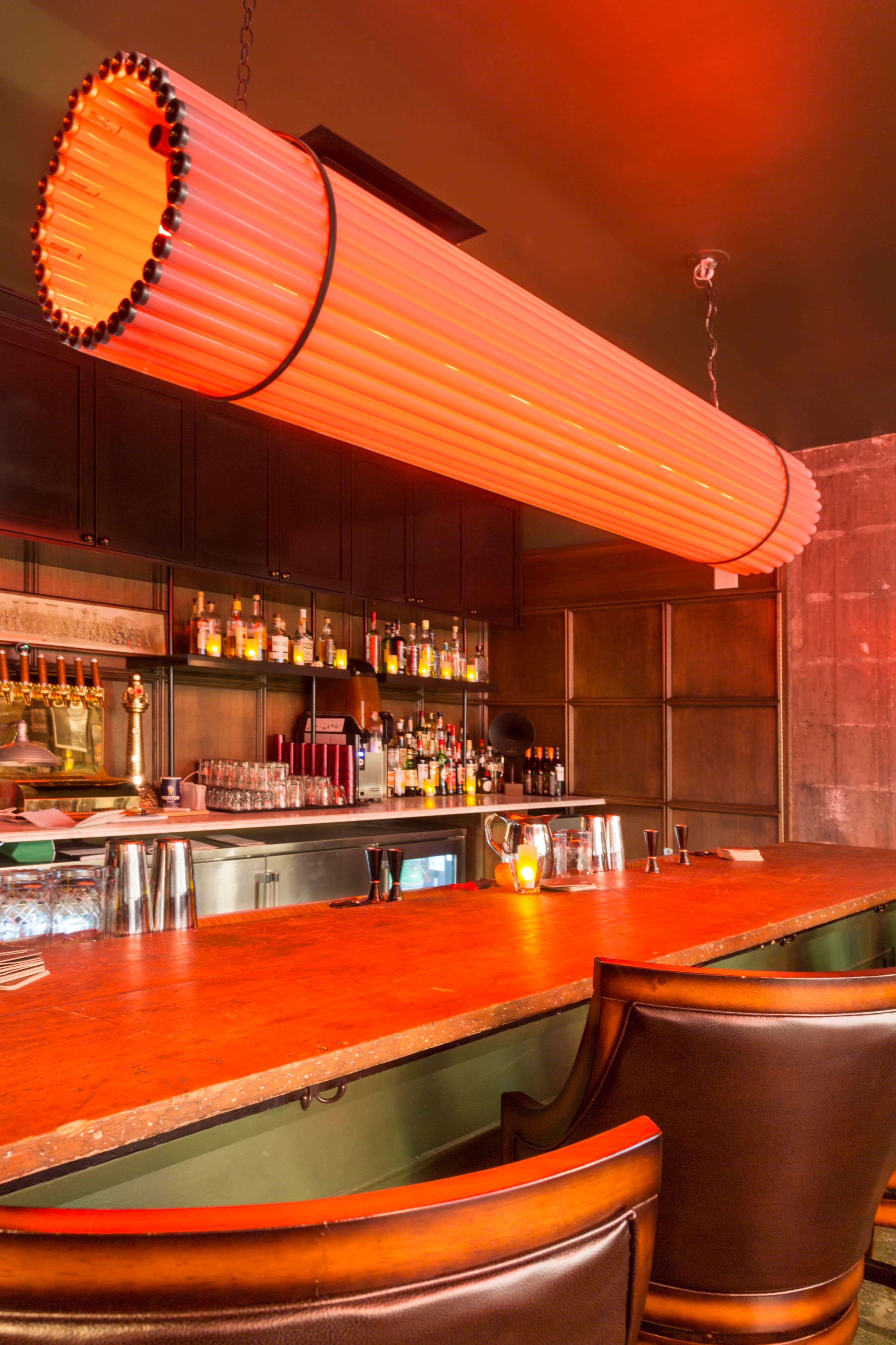 A modern bar area features a long, cylindrical pendant light above a polished wooden counter and a variety of bottles arranged on shelves behind the bar.