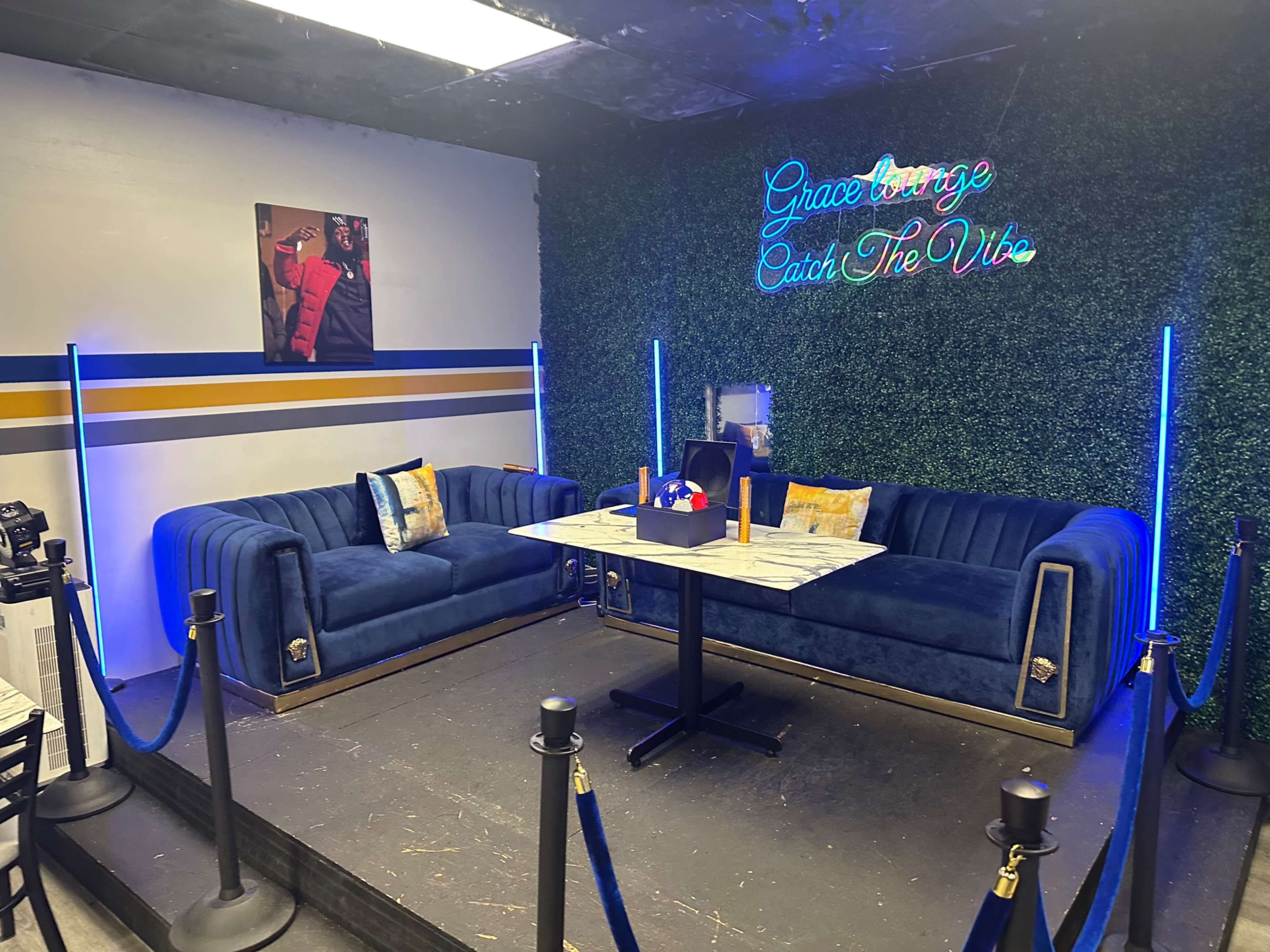 The image shows a cozy lounge area with plush blue sofas, a black table, and neon signage against a green wall.