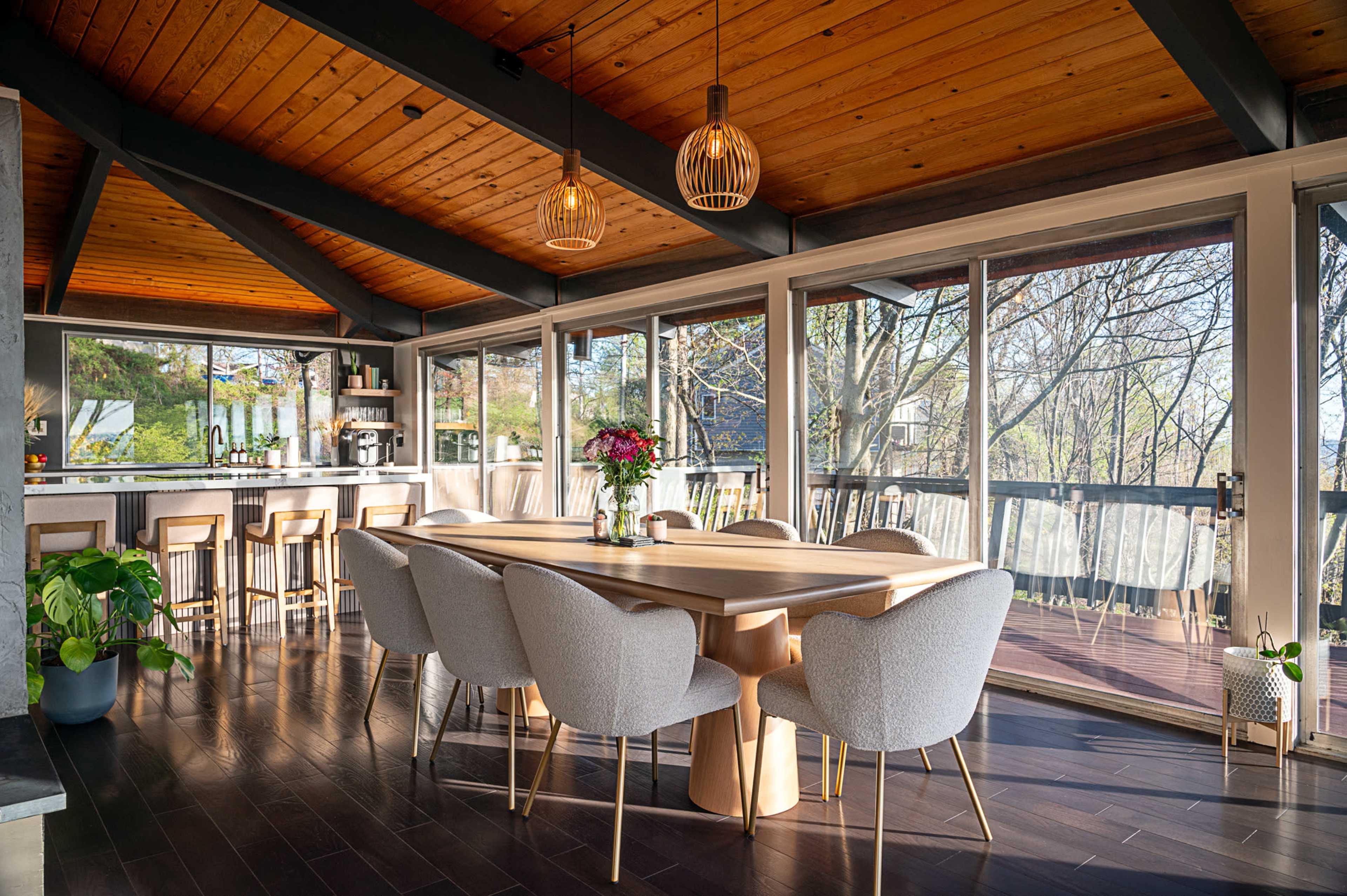 A modern dining area features a wooden table surrounded by upholstered chairs, with large glass windows that overlook a natural setting.
