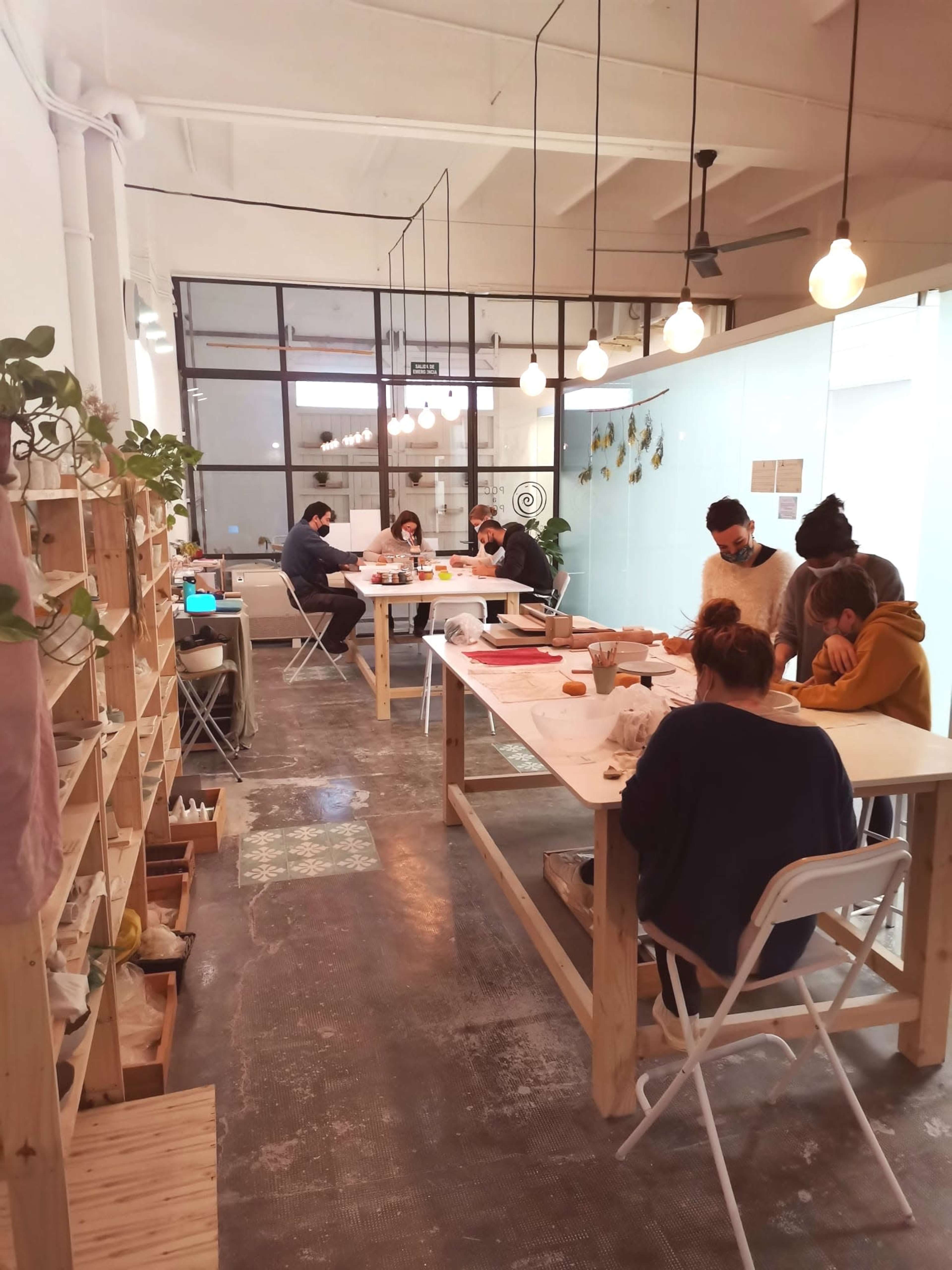 A spacious workshop with several people engaged in creative activities at wooden tables, illuminated by pendant lights.