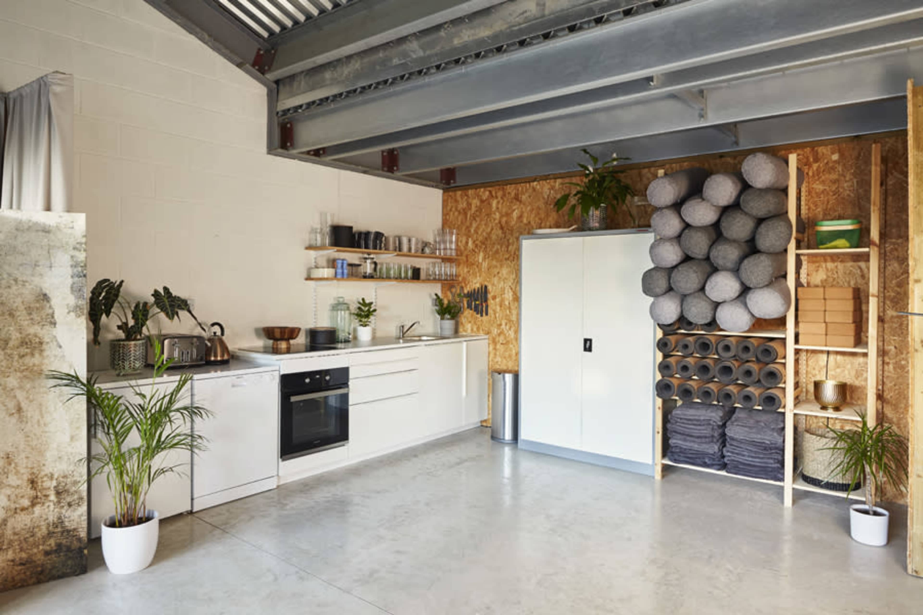 The image shows a modern kitchen space with a sleek white cabinet, various kitchenware on shelves, and a stack of exercise mats arranged on the right side.