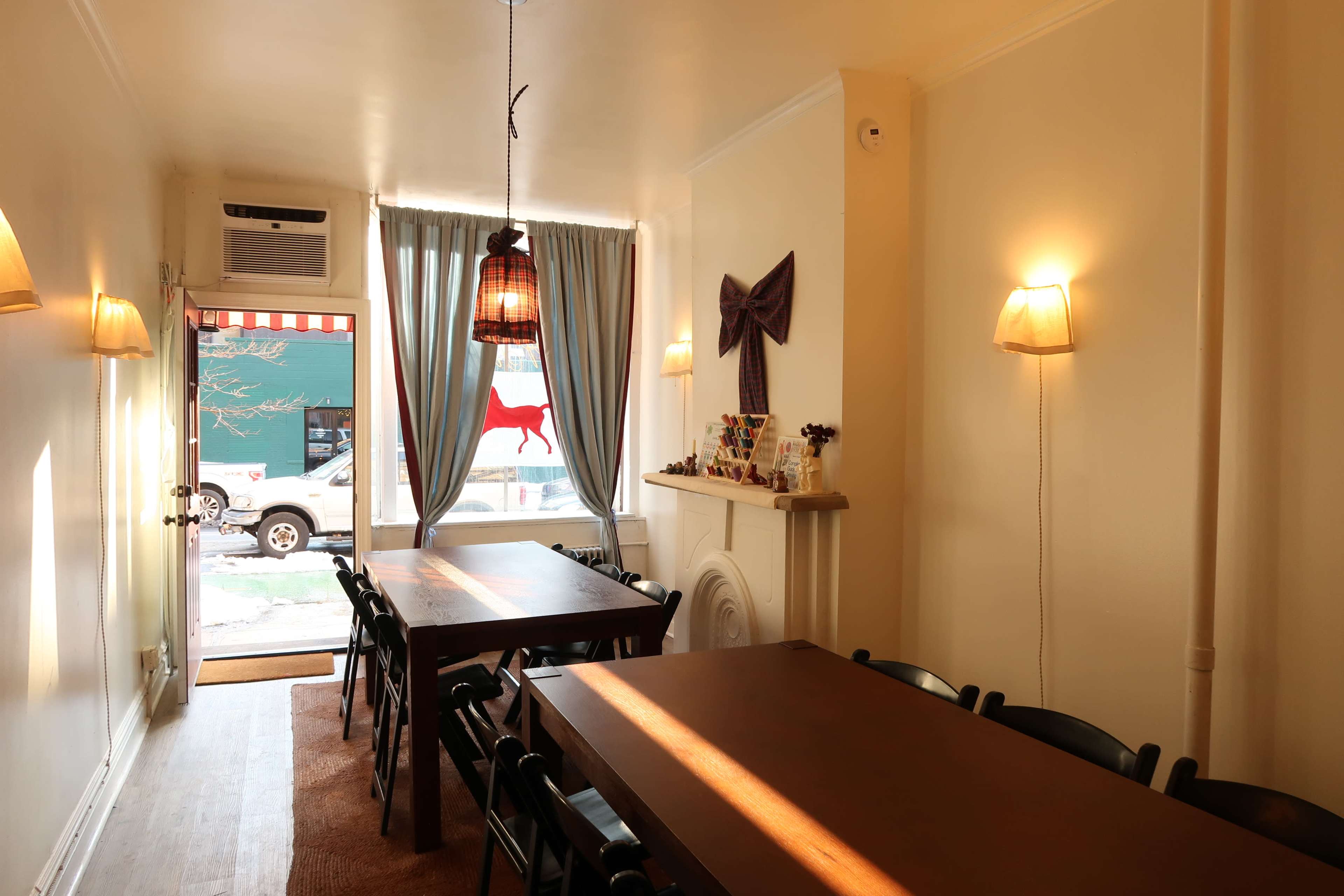 The image shows a cozy room with wooden tables, black chairs, and large windows draped with gray curtains, illuminated by warm light fixtures.