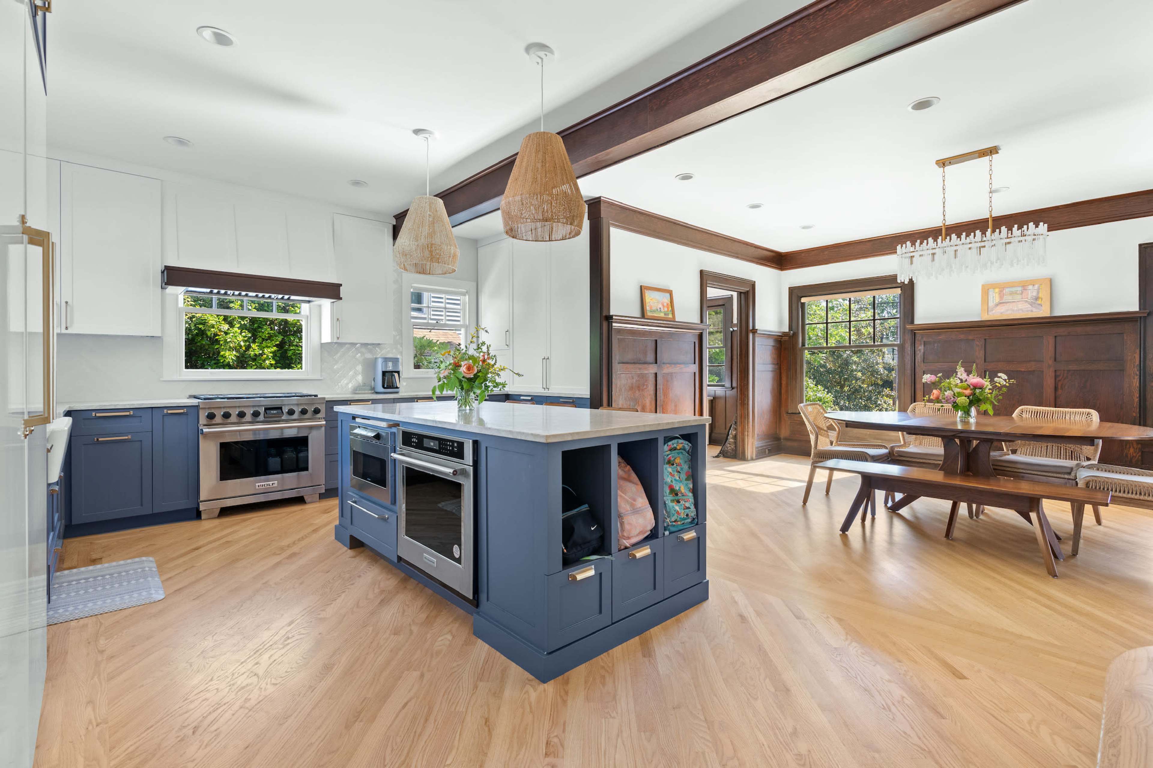 A modern kitchen with a large island, blue cabinetry, stainless steel appliances, and an adjoining dining area featuring a wooden table and natural light.