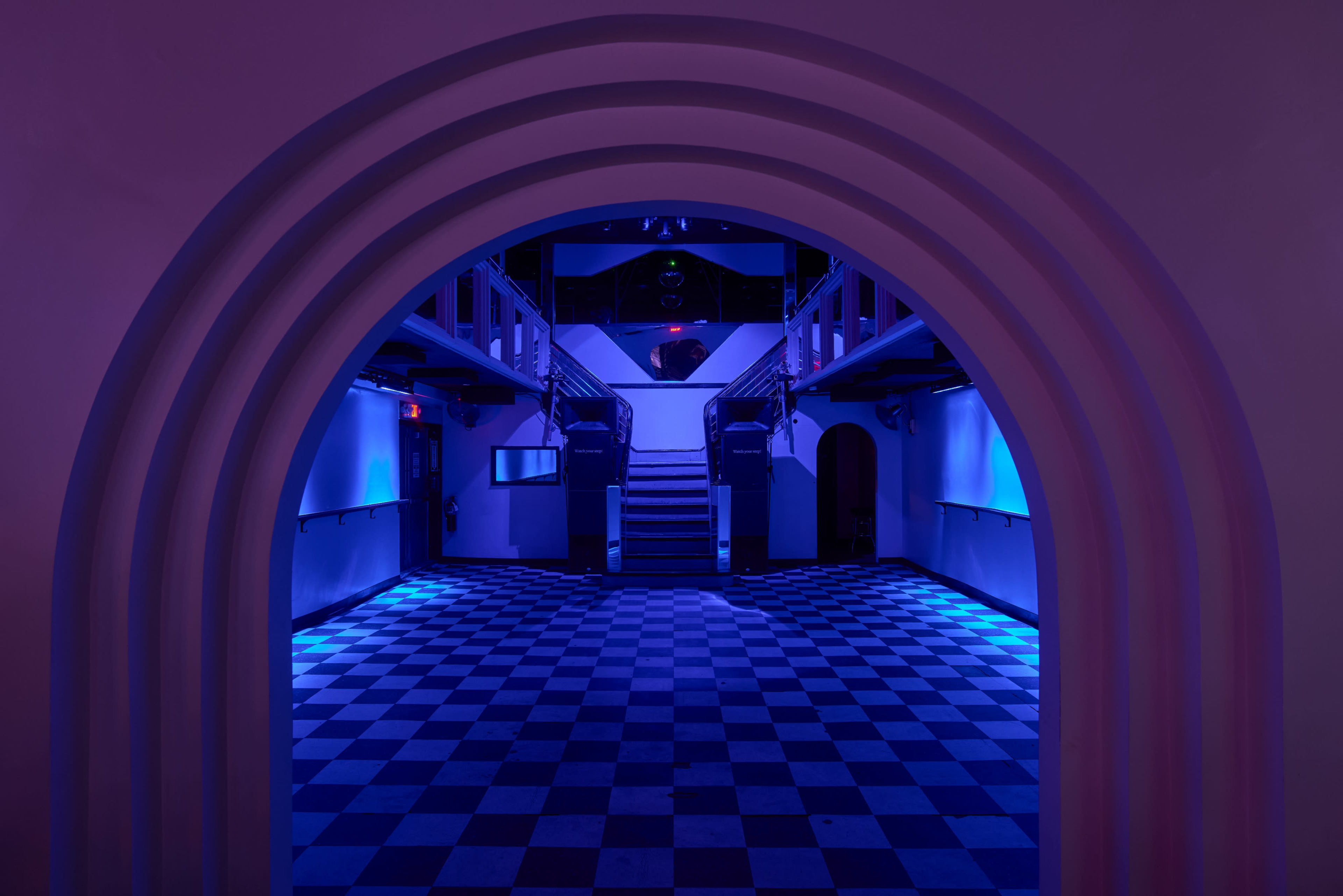 A dimly lit interior of a building with a checkered floor and an arched entrance leading to a staircase in the background.