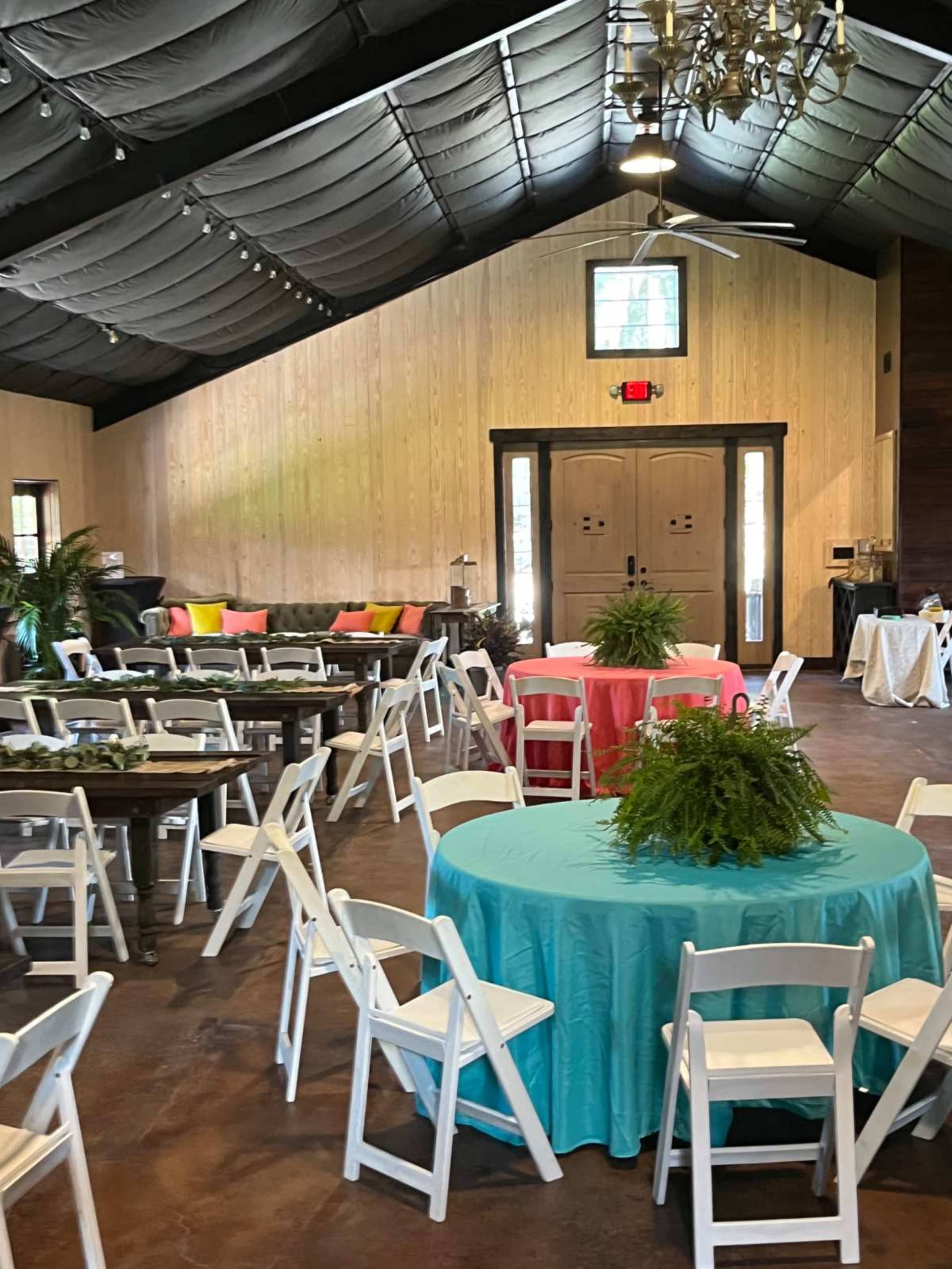 The image shows a spacious event venue with round tables covered in turquoise and pink tablecloths, surrounded by white chairs and decorated with greenery, under a high ceiling with exposed beams.