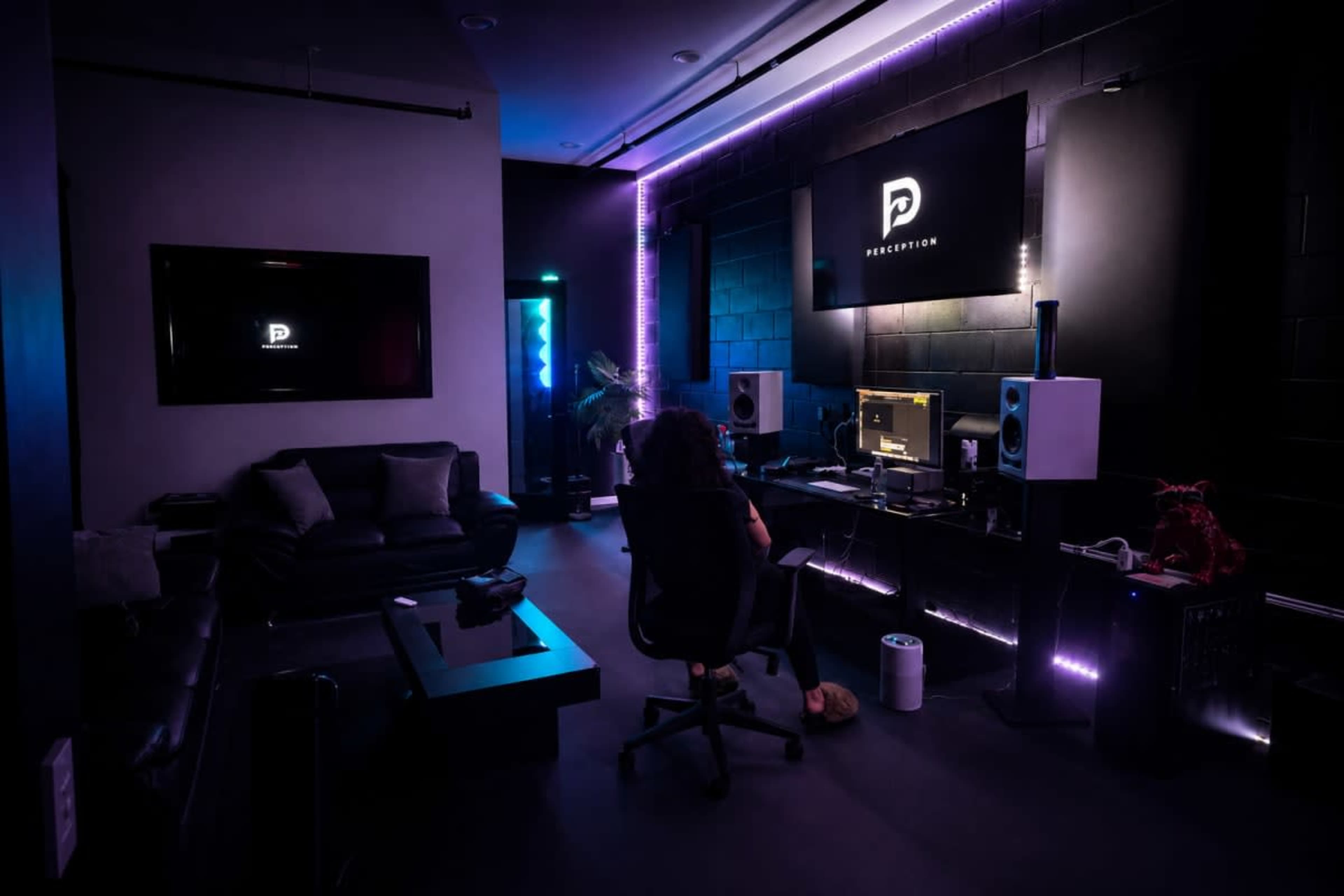A person sits in a swivel chair in a dimly lit room with modern decor, working at a desk with audio equipment and screens displaying a logo.
