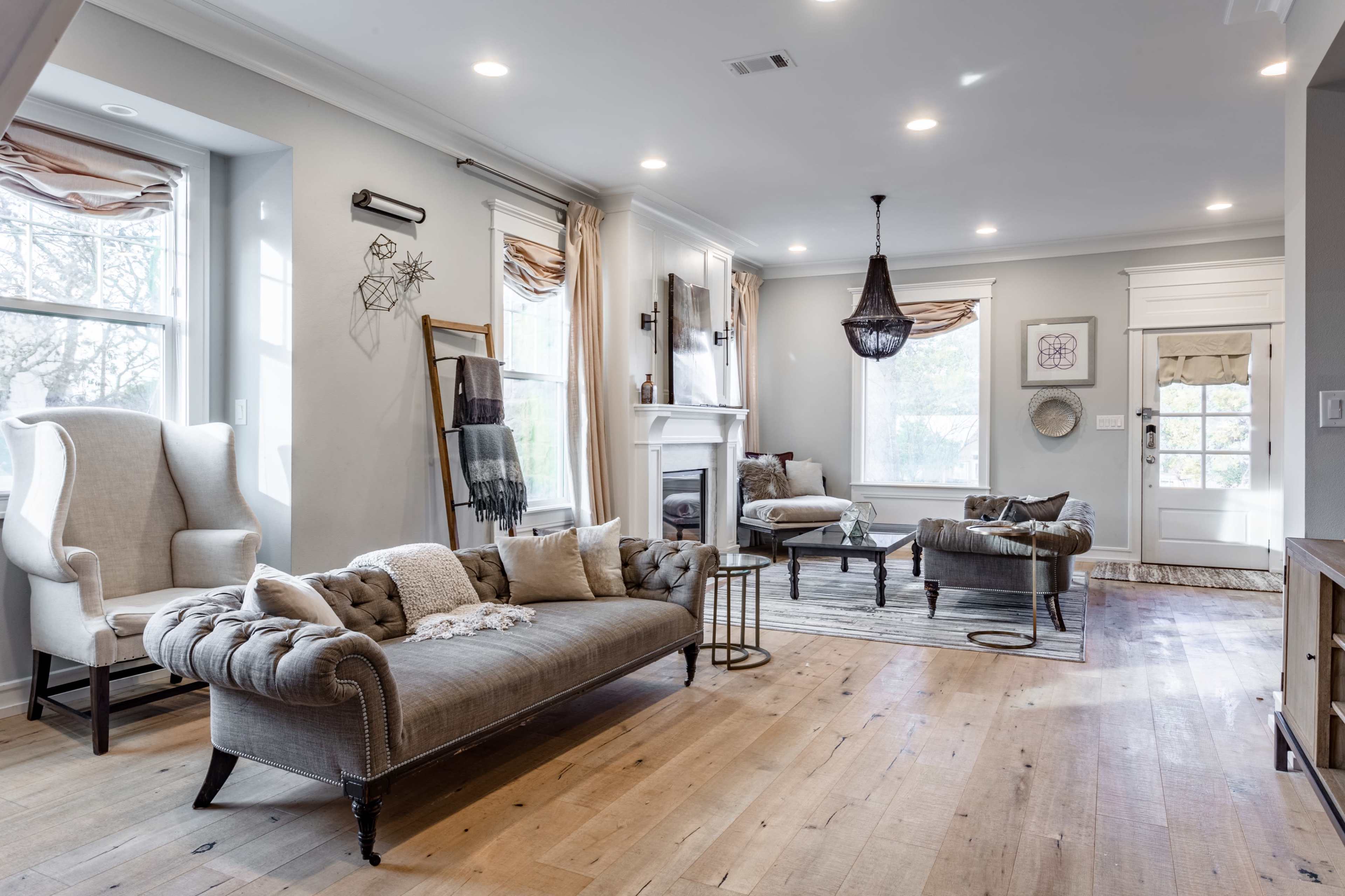 A living room features a tufted sofa, a chair, and a coffee table, all arranged around a fireplace with large windows allowing natural light.
