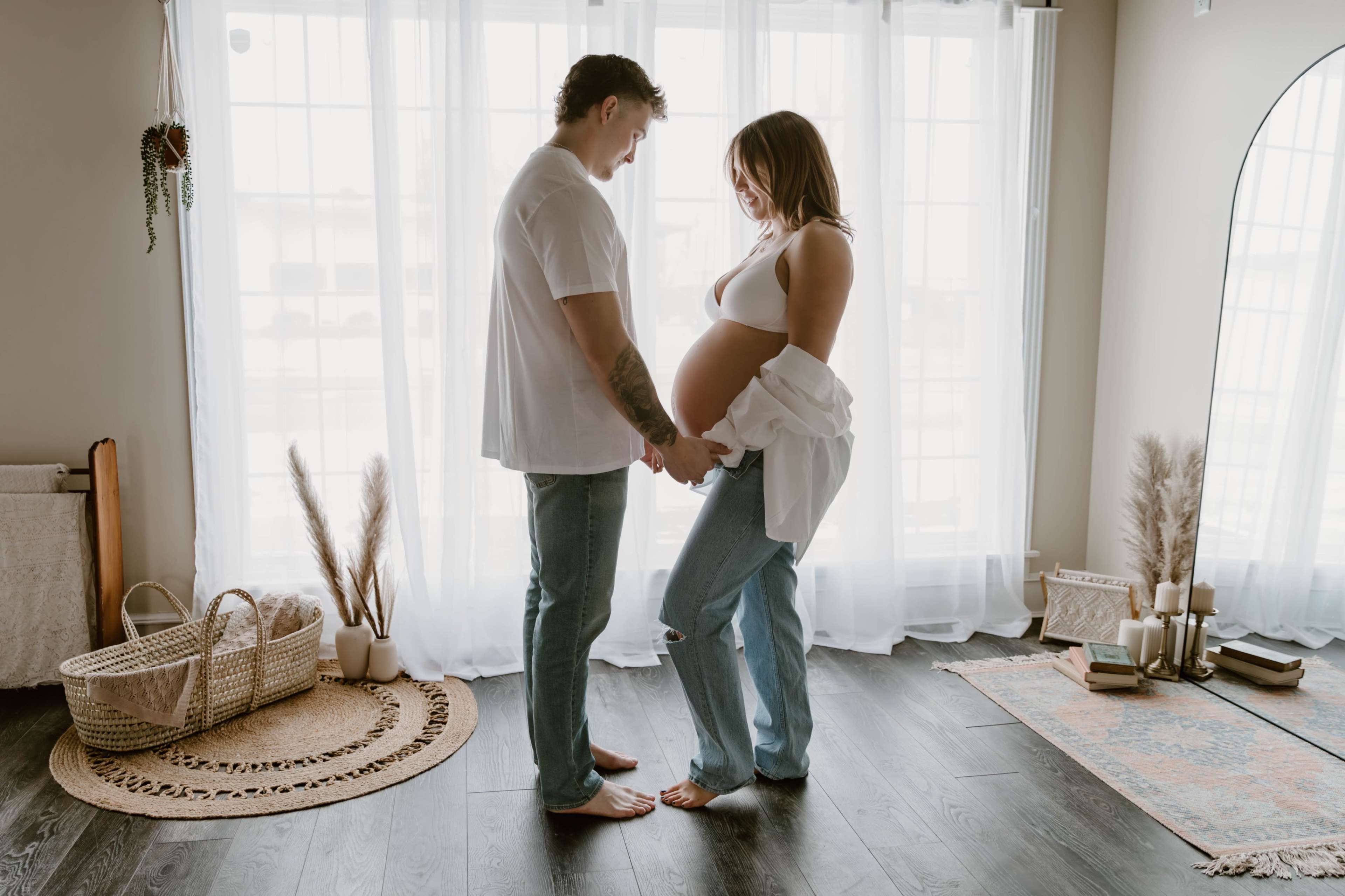 A couple stands barefoot in a well-lit room, holding hands while gazing at each other, with the woman cradling her pregnant belly.