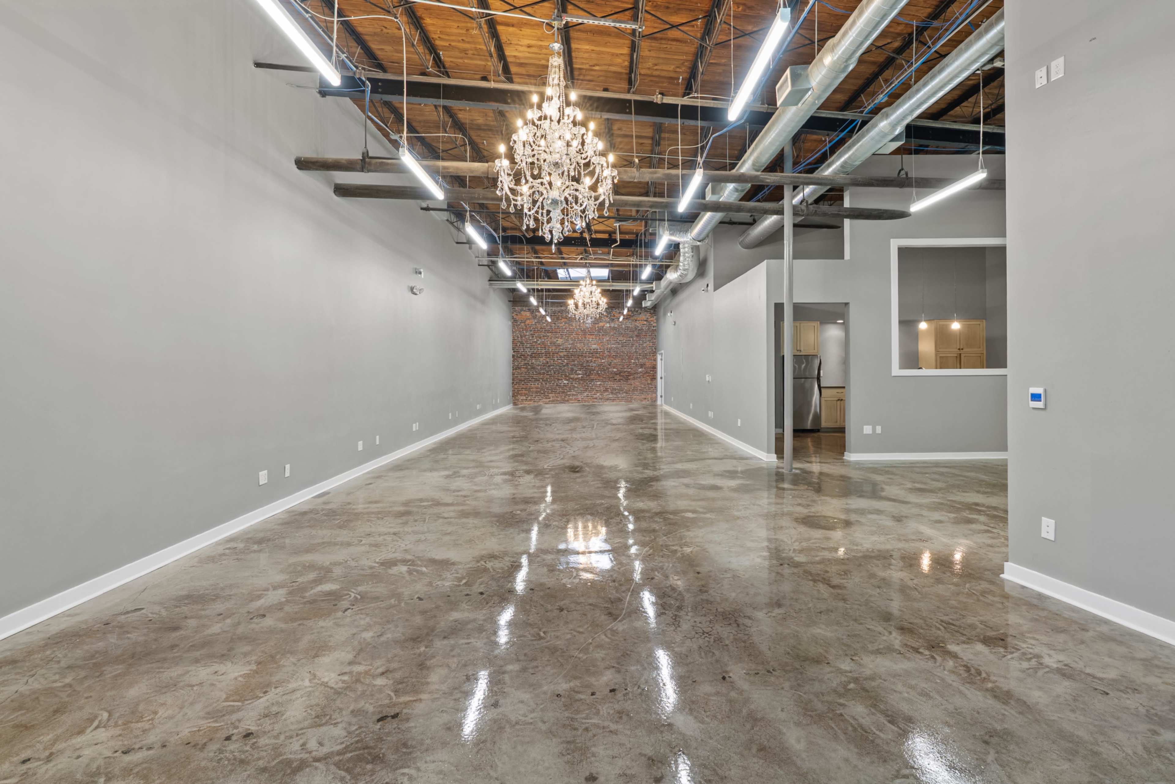 The image shows a spacious commercial interior with polished concrete floors, exposed wooden beams, and decorative chandeliers, along with a brick wall in the background.
